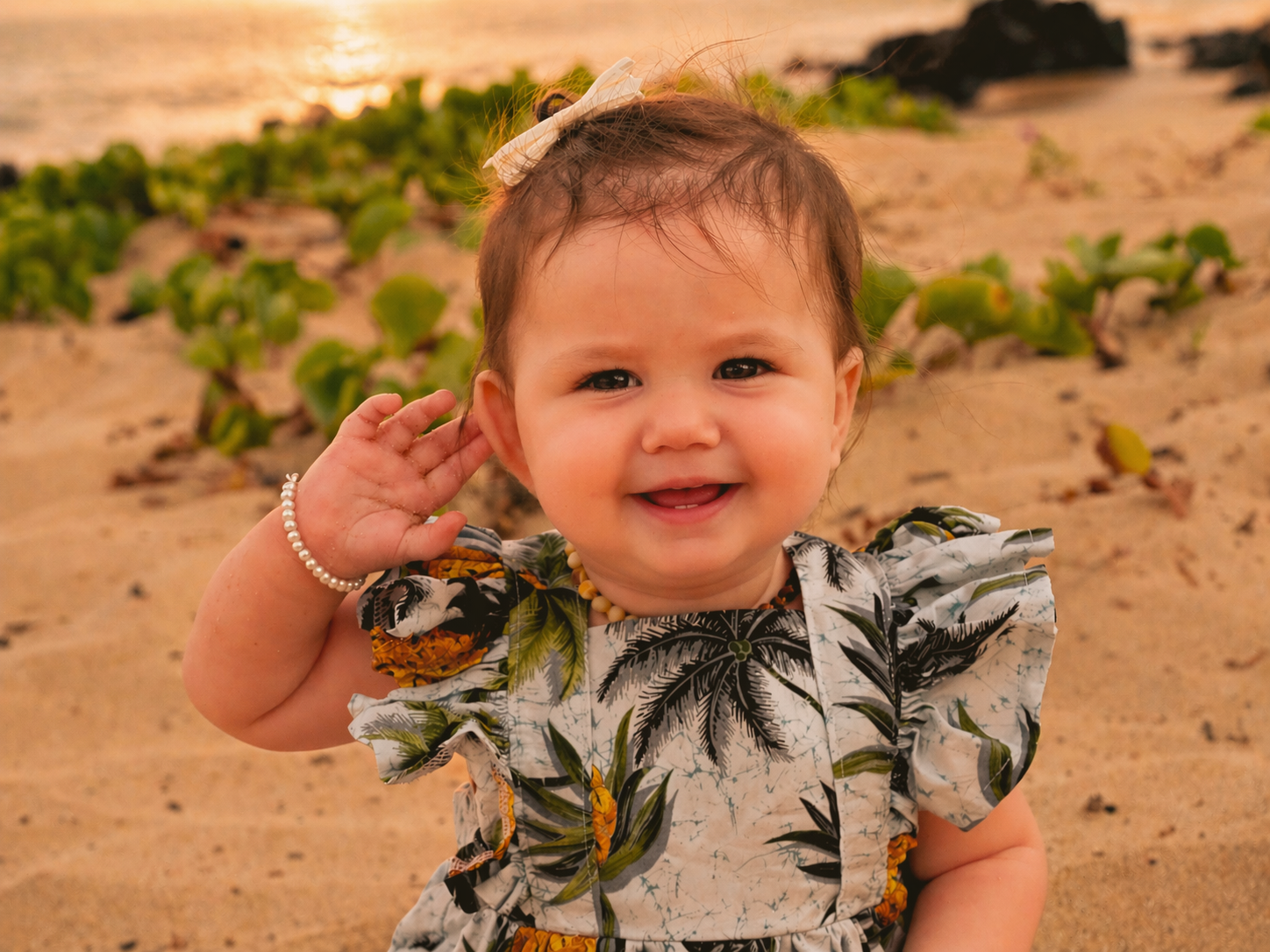 A smiling toddler girl with brown hair, wearing a patterned dress with tropical leaves and flowers, standing on the beach near the water at sunset, holding her hand to her ear.