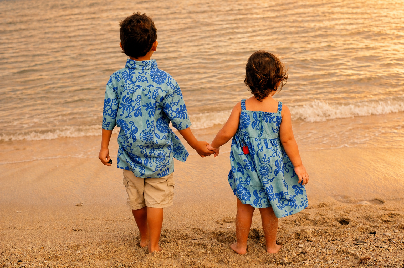 Two children, a boy and a girl, holding hands on the beach at sunset, facing the ocean, with sand and gentle waves in the background.