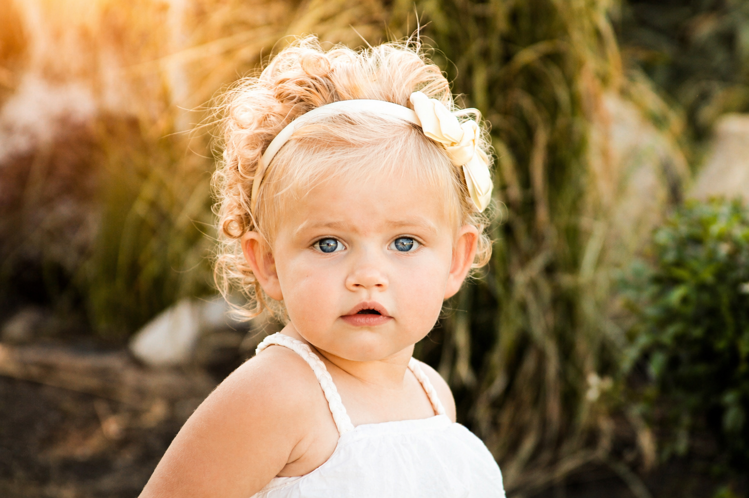 A young girl with curly blonde hair, blue eyes, and a white headband with a bow, wearing a white sleeveless dress, standing outdoors with a blurred natural background.