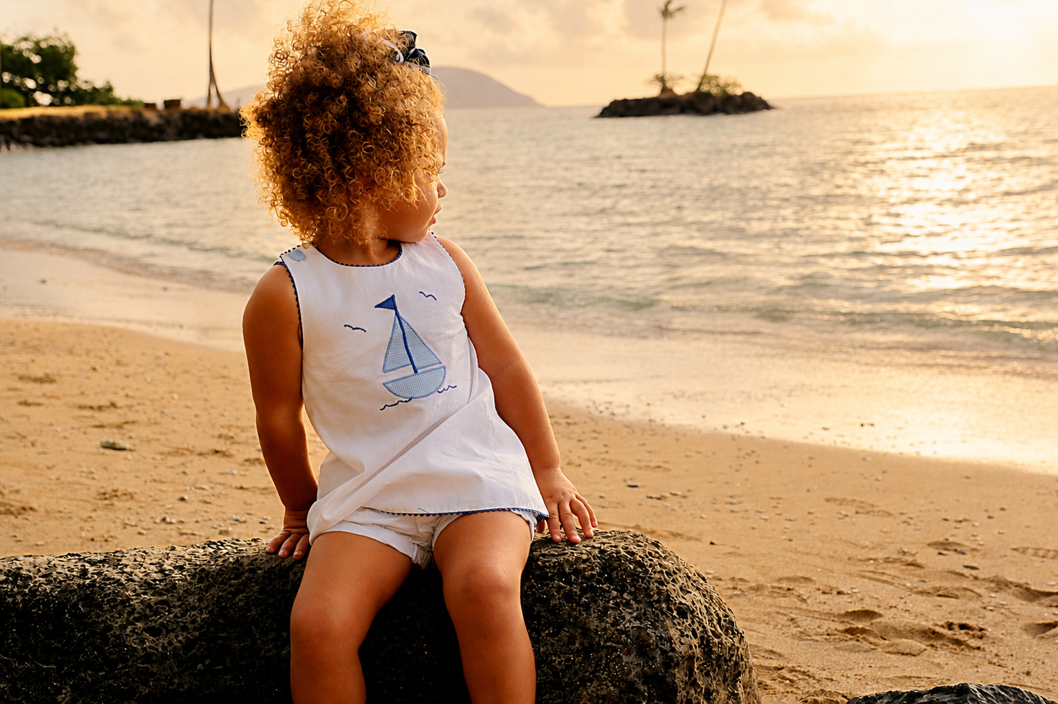 A young girl with curly hair sitting on a rock at the beach during sunset, looking toward the water with small islands and palm trees in the background.