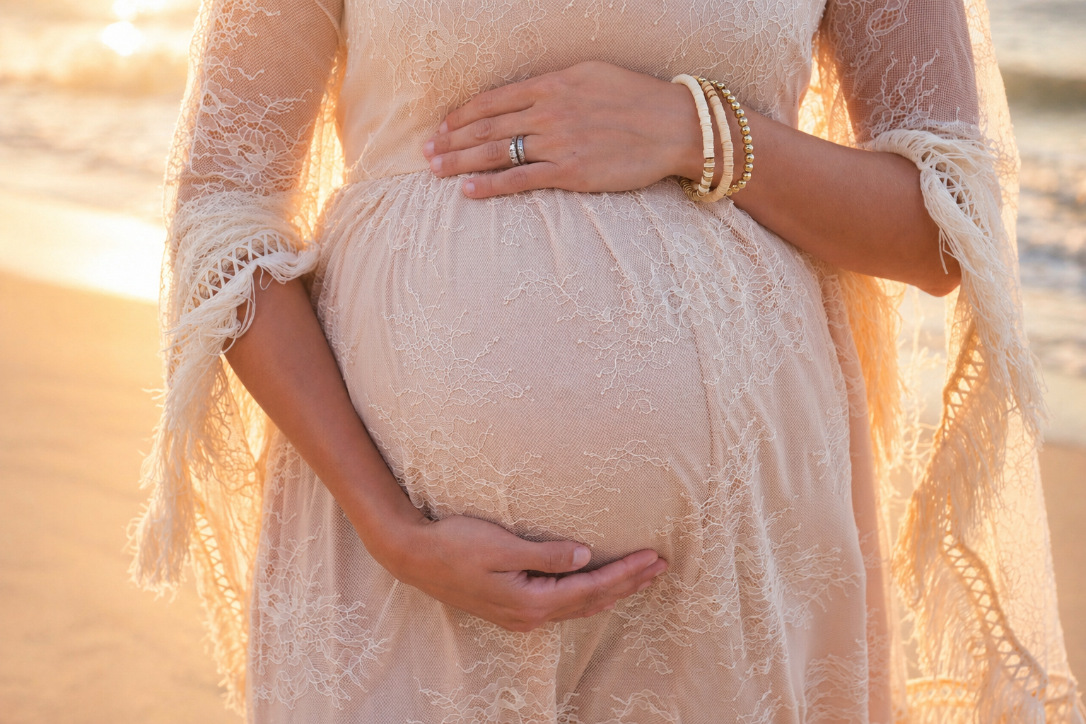 Close-up of a pregnant woman's belly at the beach during sunset, with her hands gently cradling her belly and wearing a lace dress and jewelry.