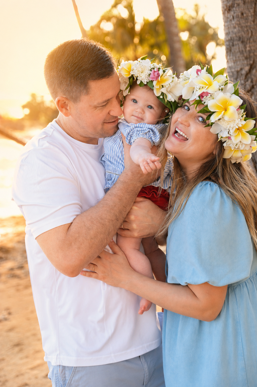 Family of three at the beach during sunset, with two adults holding a baby who is wearing a flower crown and a blue striped outfit.