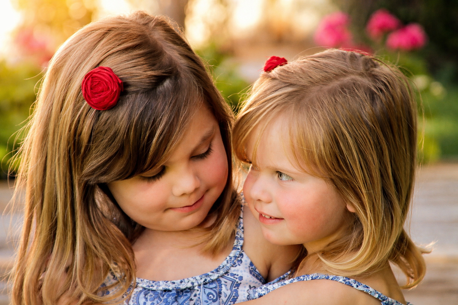 Two young girls with red flower hair accessories hugging outdoors in sunlight, with pink flowers and greenery in the background.
