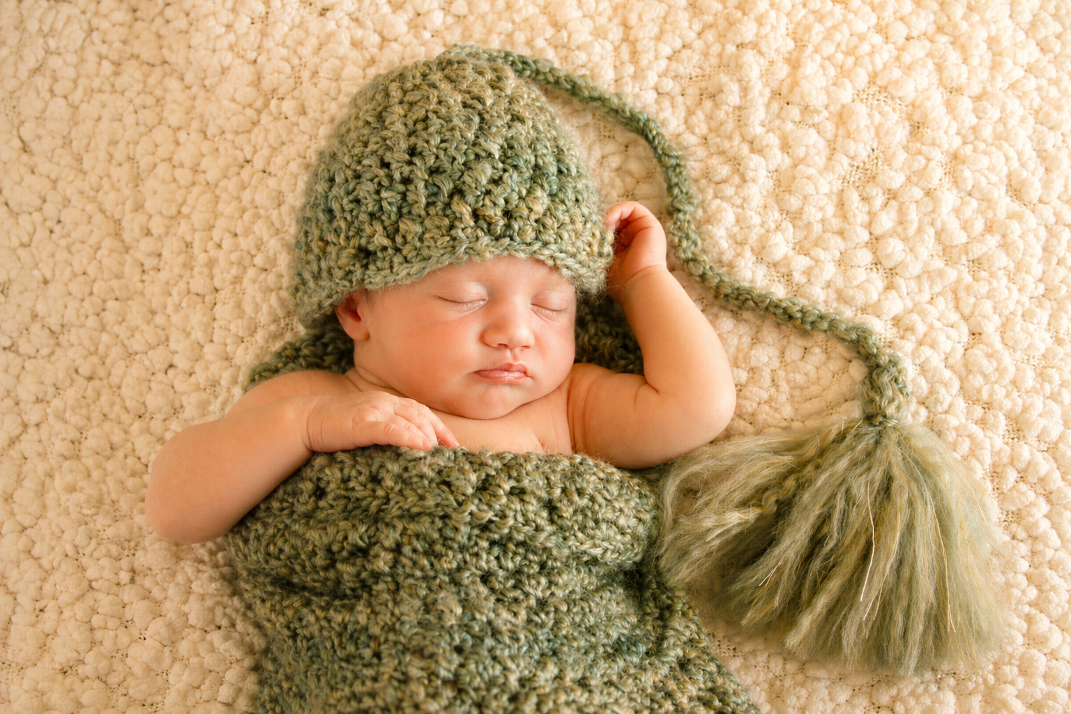 A sleeping baby wrapped in a chunky knit blanket, wearing a matching knit hat with a tassel. The baby is lying on a textured, cream-colored blanket.