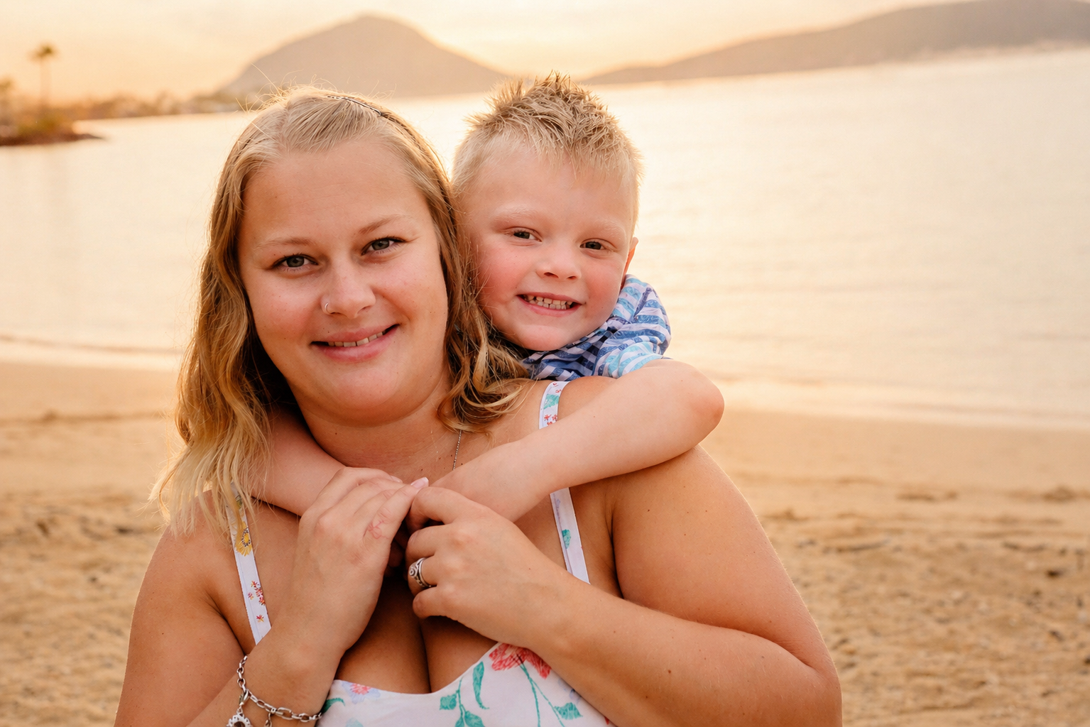 A woman with blonde hair and a young boy with blonde spiky hair smiling at the camera, outdoors on a beach during sunset with water and mountains in the background.