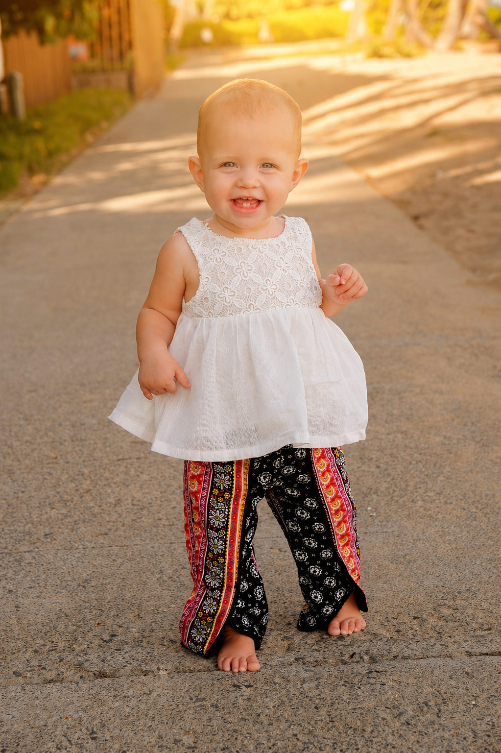 A young girl smiling and walking barefoot on a paved path during golden hour. She is wearing a white lace top with a flowy skirt and black, red, and yellow patterned pants. The background features a sunny, outdoor setting with trees and a wooden fenc