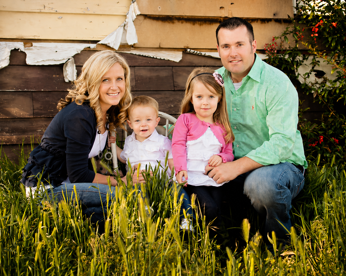 A family of four sitting in a grassy garden in front of a weathered wooden shed, smiling at the camera. The mother has blonde curls, the father has dark hair, the boy has short light hair, and the girl has long light brown hair with a pink bow