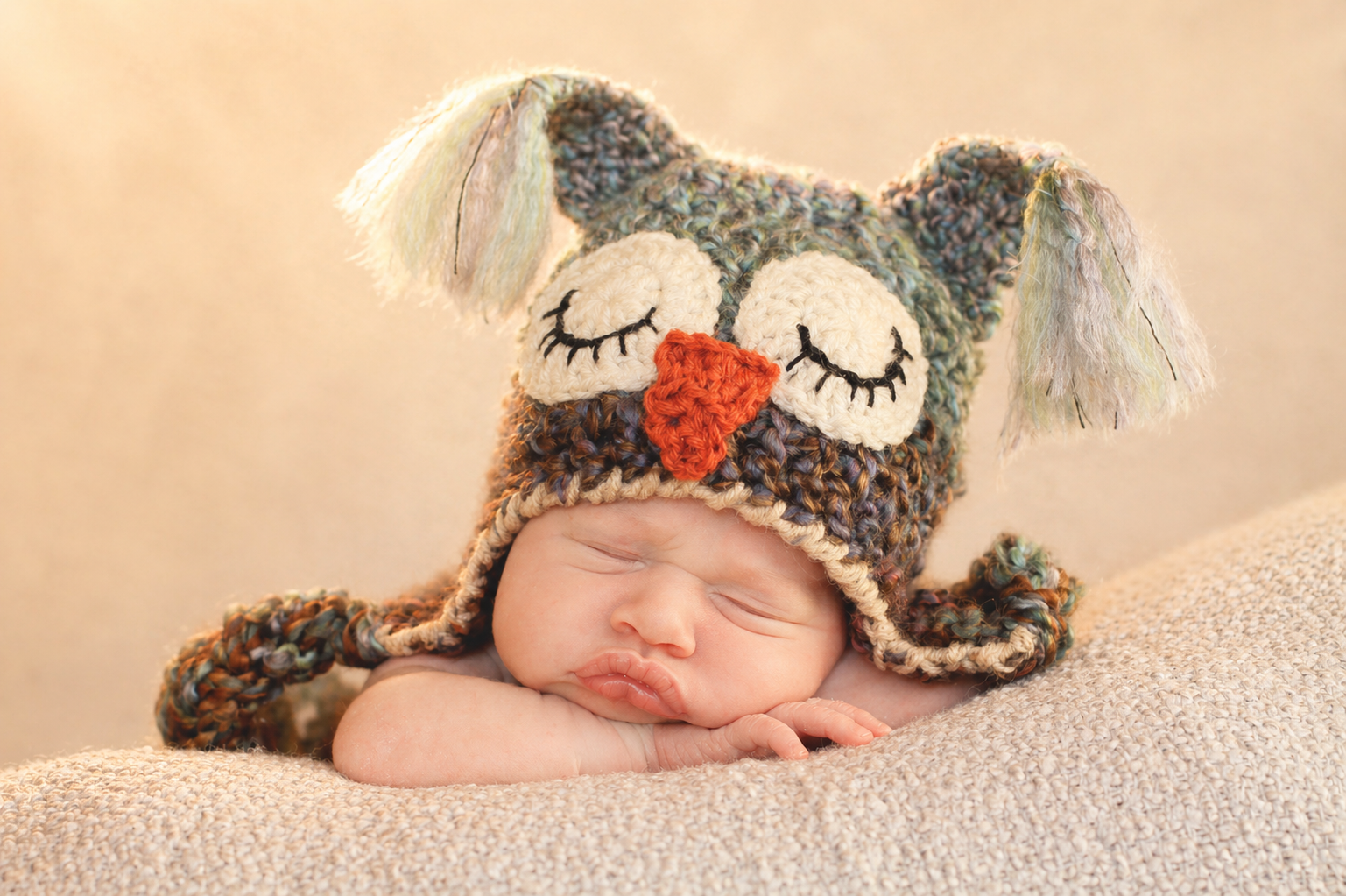 A sleeping baby resting on a soft beige blanket, wearing a knitted owl hat with closed eyes, an orange beak, and floppy ears.