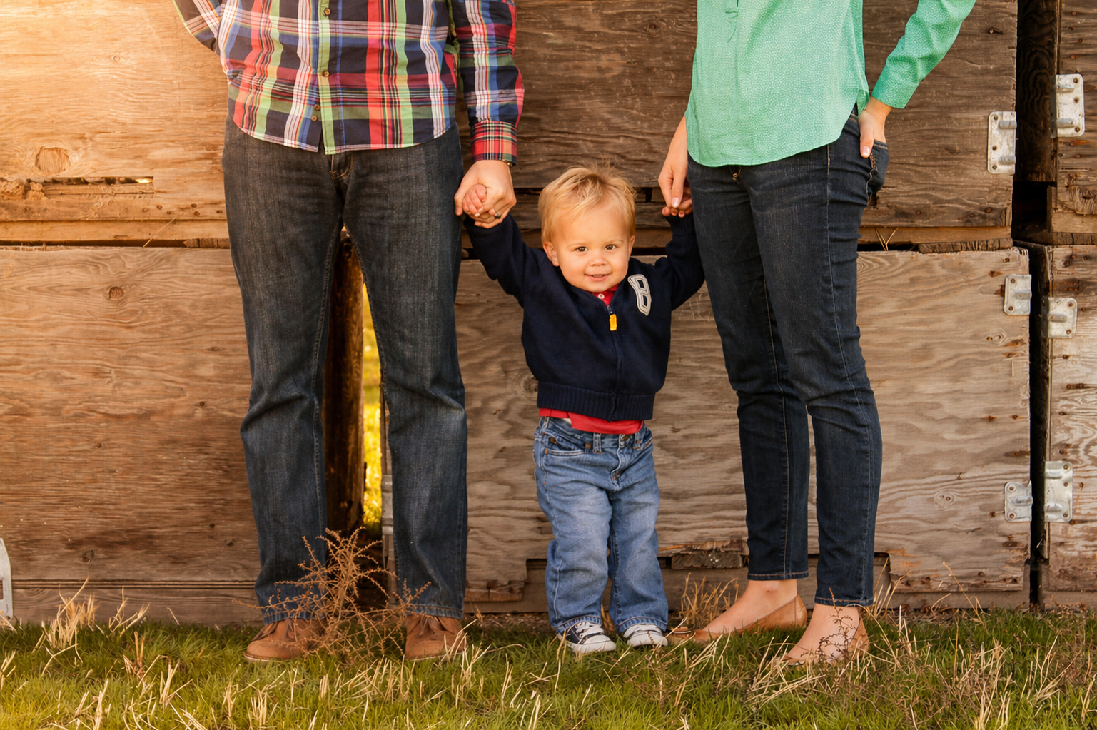 A young boy holding hands with two adults, standing on grass in front of a wooden wall.