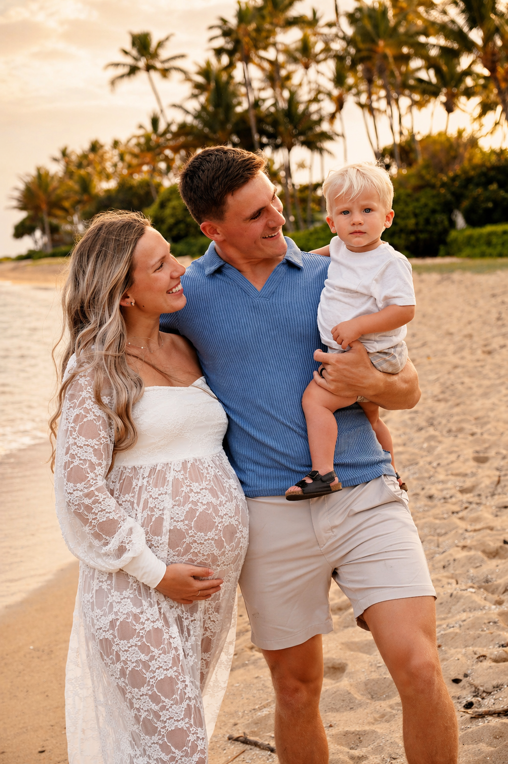 A smiling family walking on the beach at sunset, including a pregnant woman, a man holding a young boy, with palm trees in the background.