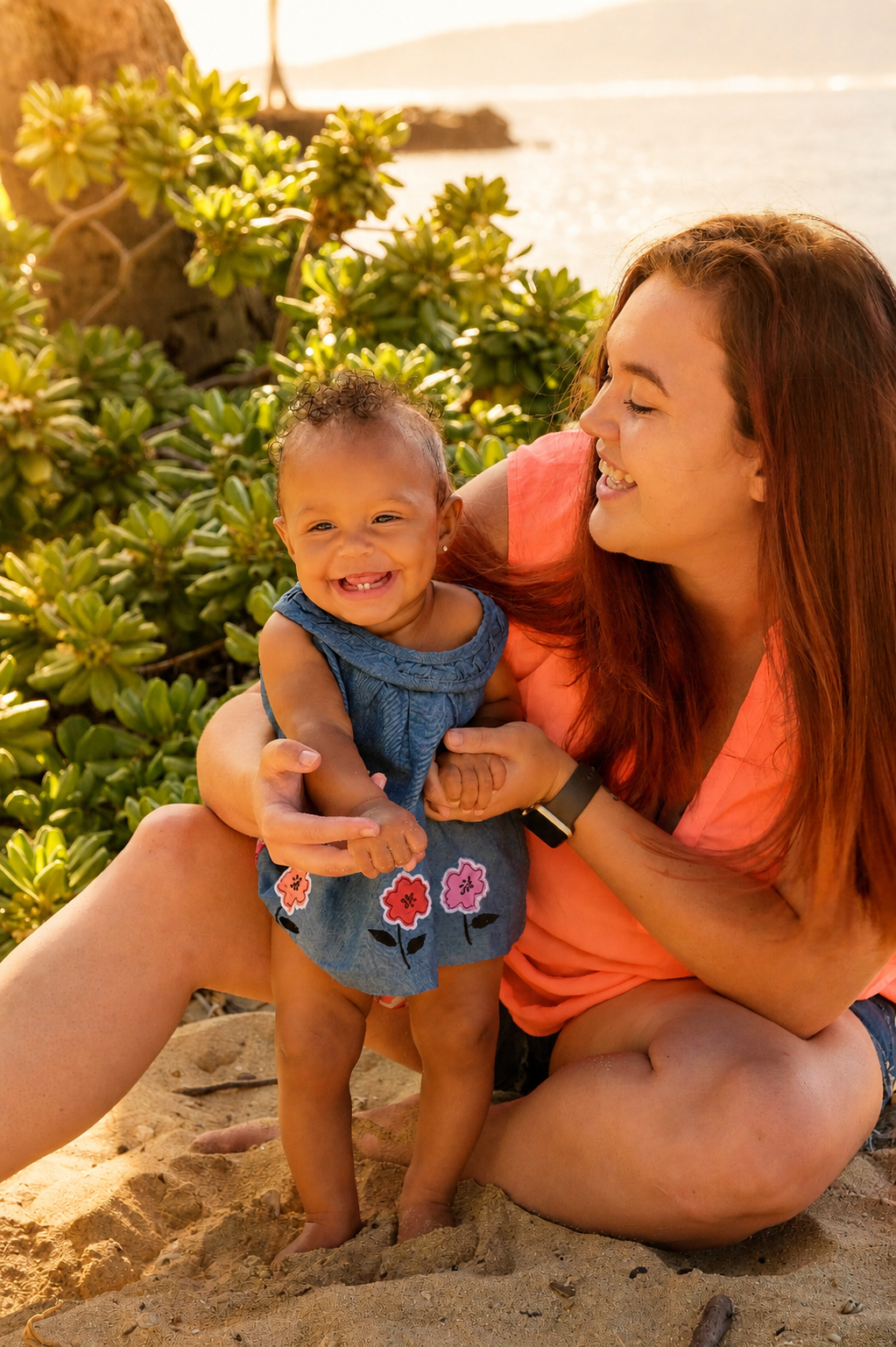 A woman and a young girl smiling and playing on a sandy beach near green plants with the ocean in the background.