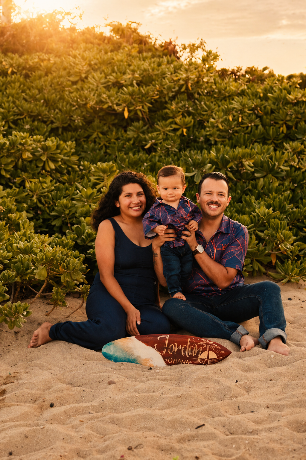 A family of three sitting on the sand at the beach during sunset, with green bushes in the background and a surfboard with 'Jordan Oahu' written on it in front of them.