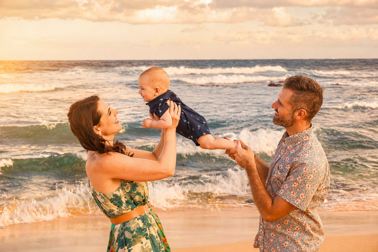 A family of three enjoying time together at the beach at sunset. The woman is holding up a smiling baby girl, and the man is holding the baby's feet, both smiling at each other.