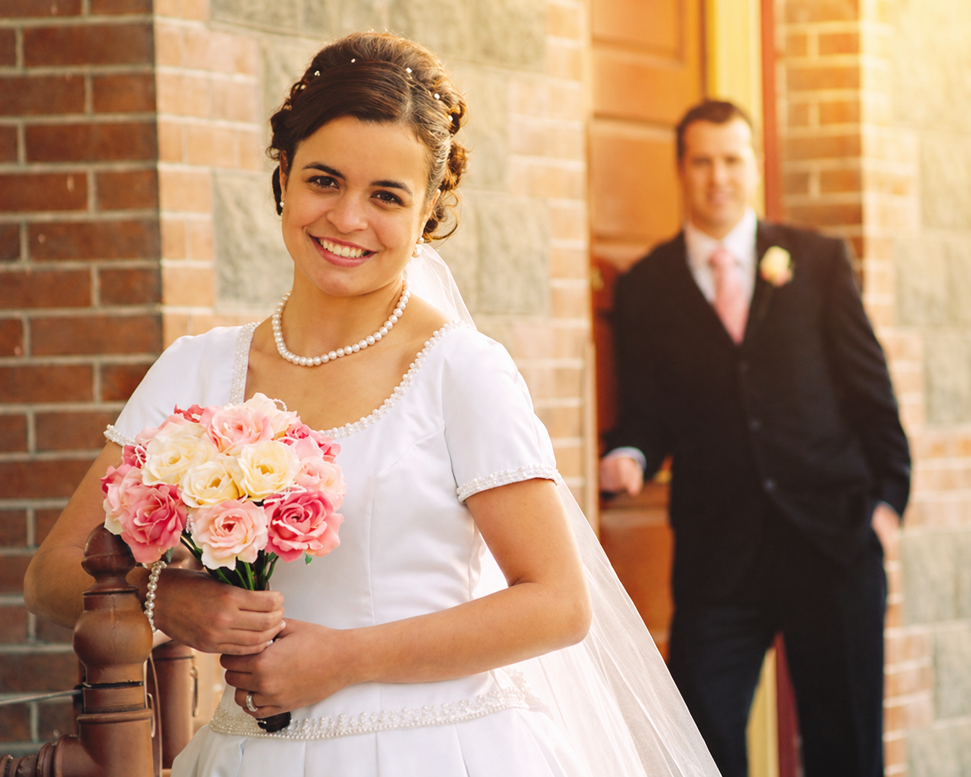 A smiling bride holding a bouquet of pink and cream roses, standing in front of a brick wall with a groom in a black suit in the background.
