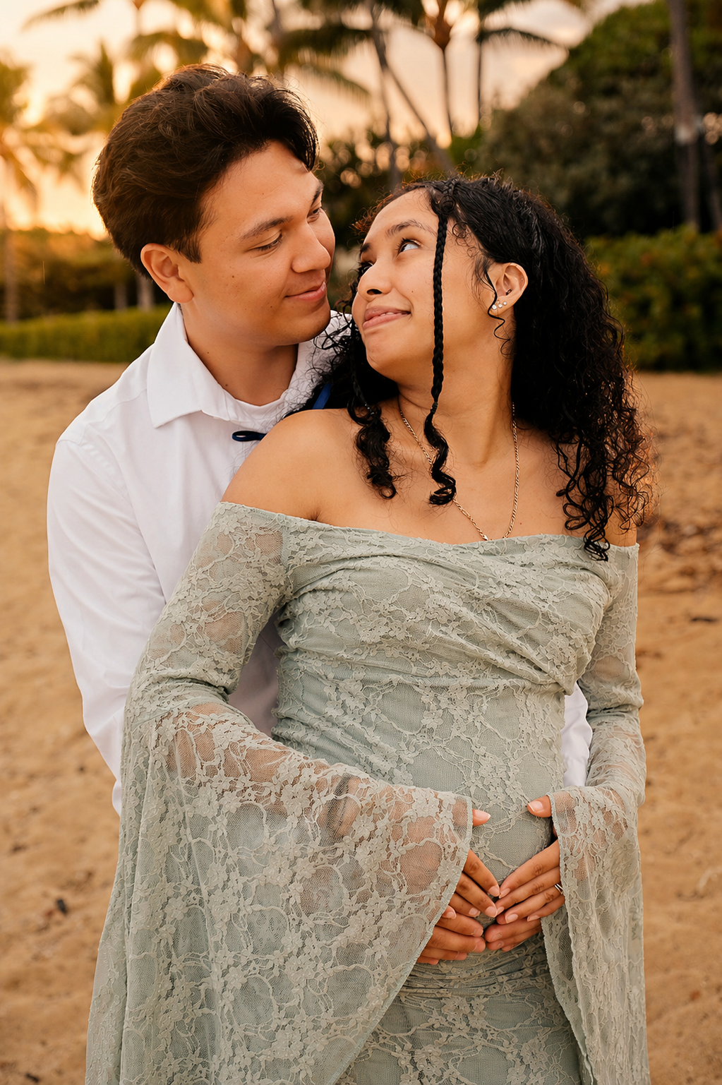 A couple stands on a sandy beach with palm trees in the background during sunset, embracing and gazing into each other's eyes.