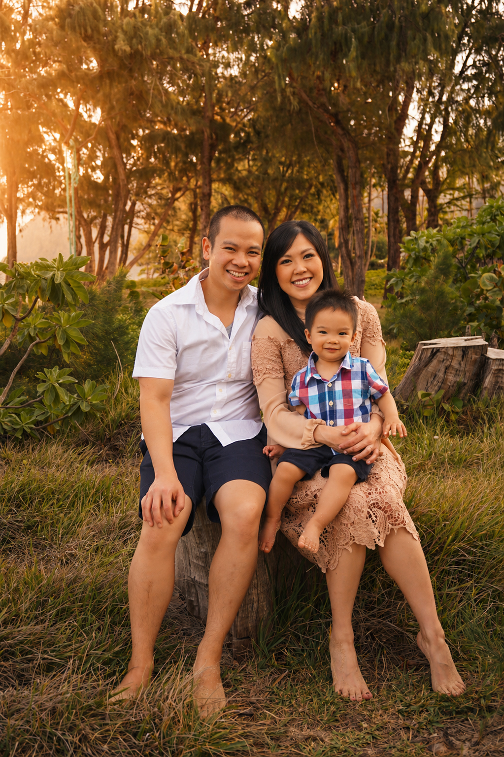 A family of three sitting on a log outdoors in a grassy area with trees at sunset, smiling at the camera.