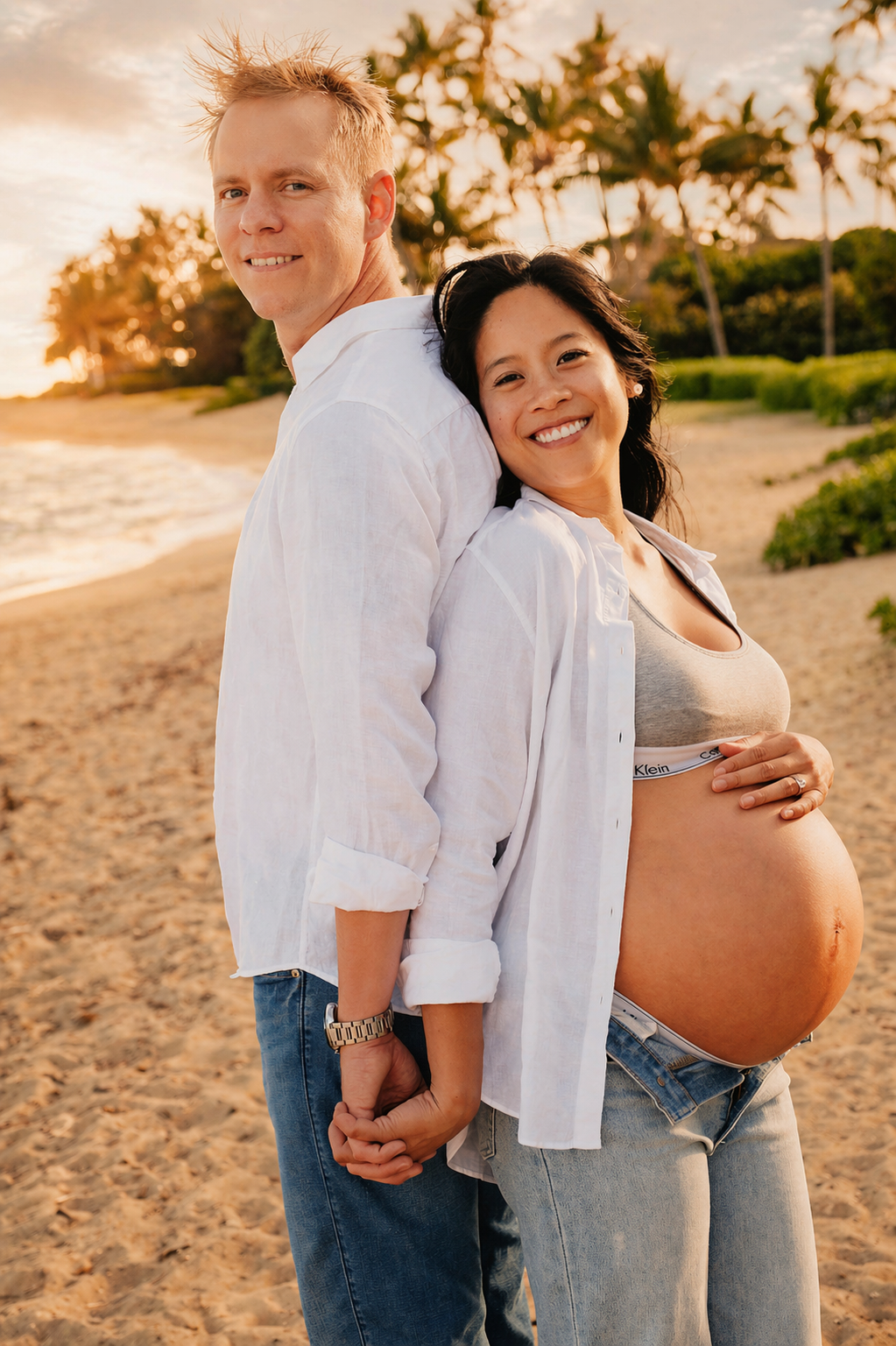 A smiling pregnant woman in a beige sports bra and open white shirt, with long dark hair, standing on a sandy beach with palm trees in the background, holding her belly. Beside her, a man in a white shirt and blue jeans, smiling and holding her hand,