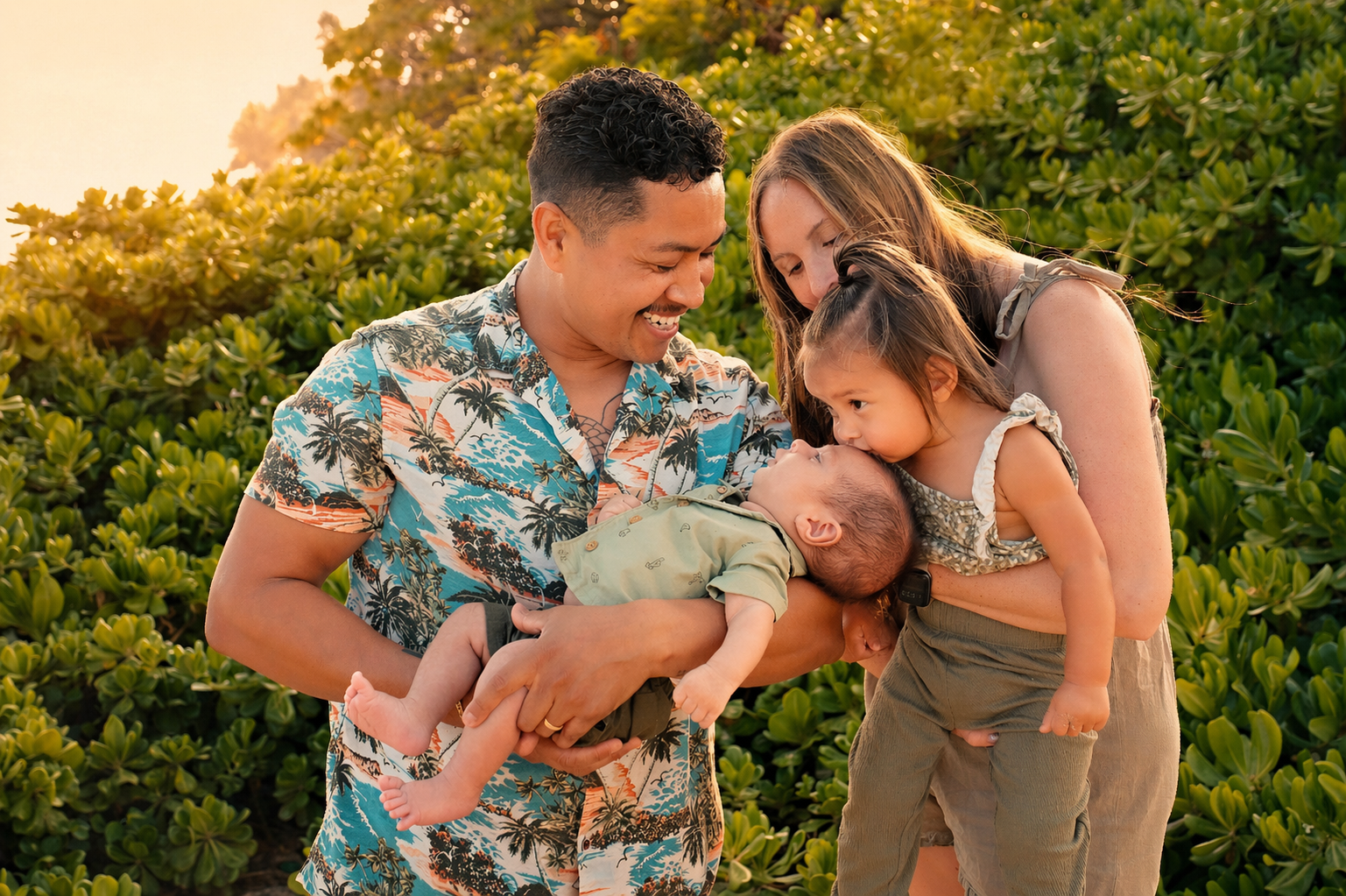 A family of four in a lush green outdoor setting during sunset, with the father holding a baby, the mother leaning in to kiss a young girl, all smiling warmly.