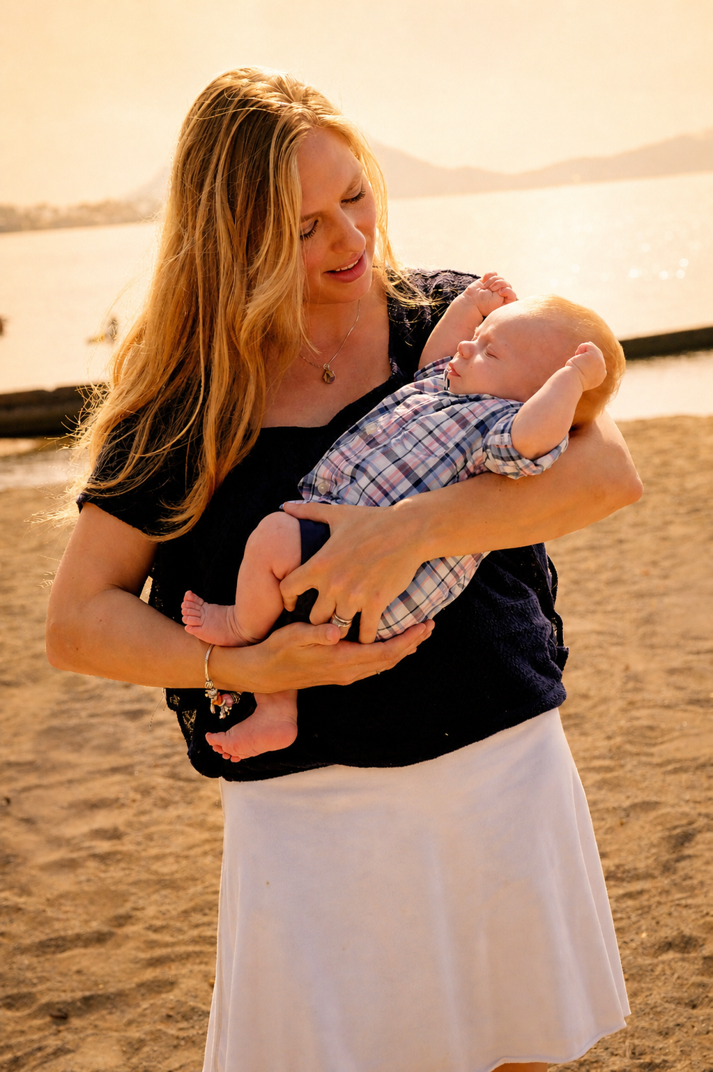 A woman with long blonde hair holding a baby with red hair on a beach at sunset