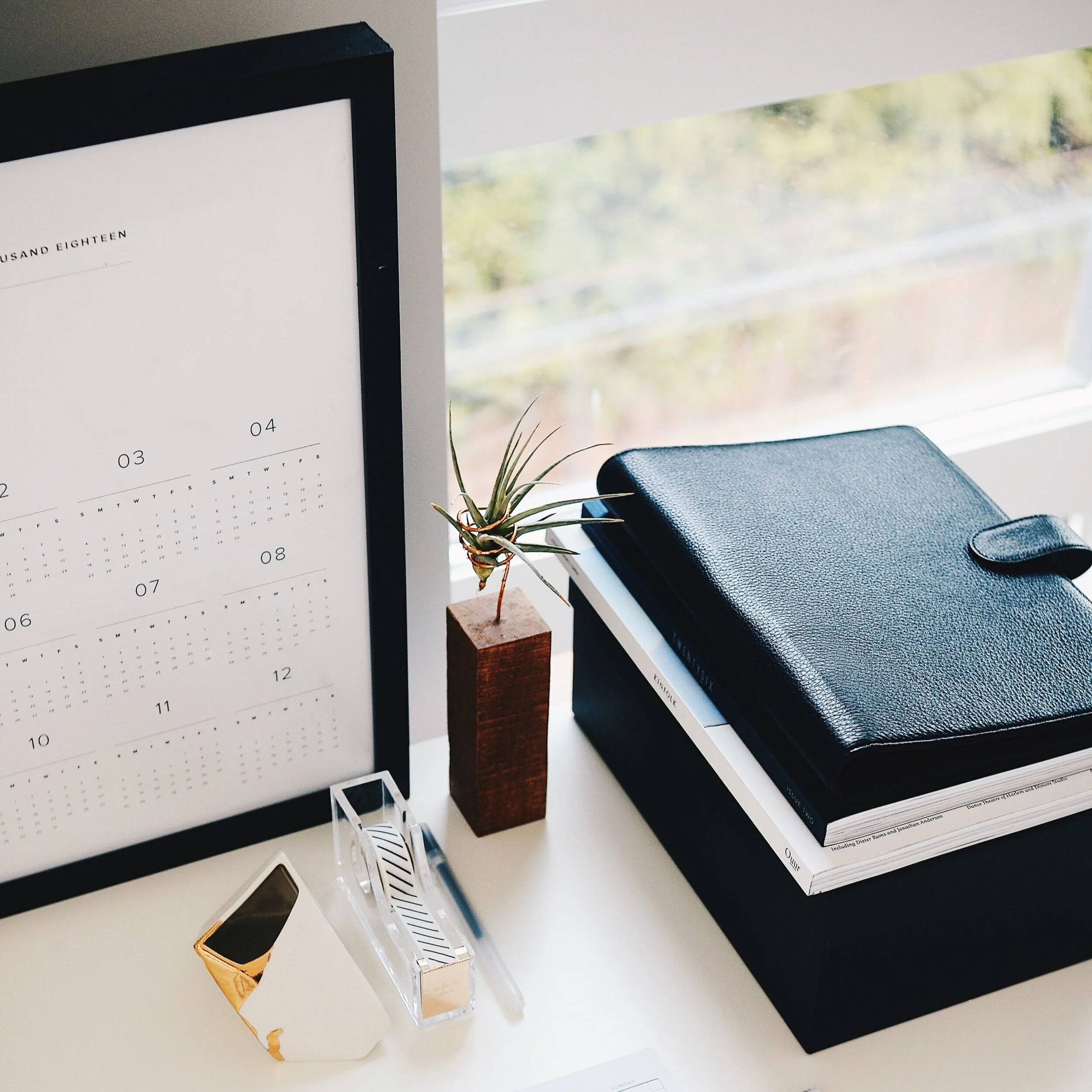 Desk with a computer monitor showing a calendar page, a potted plant, a closed black leather notebook, a smartphone in a decorative holder, and several clear acrylic stands with rings on a white surface near a window with a view of trees and shrubs outside.