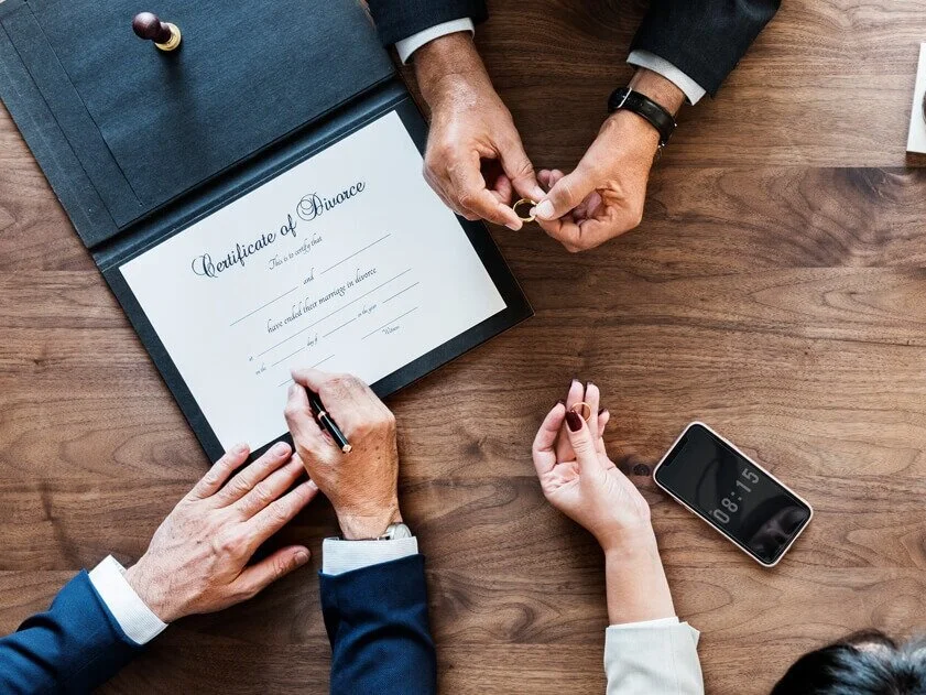A person signing a divorce certificate on a wooden table, with a hand receiving a wedding ring, a smartphone displaying the time 08:15, and other hands visible.