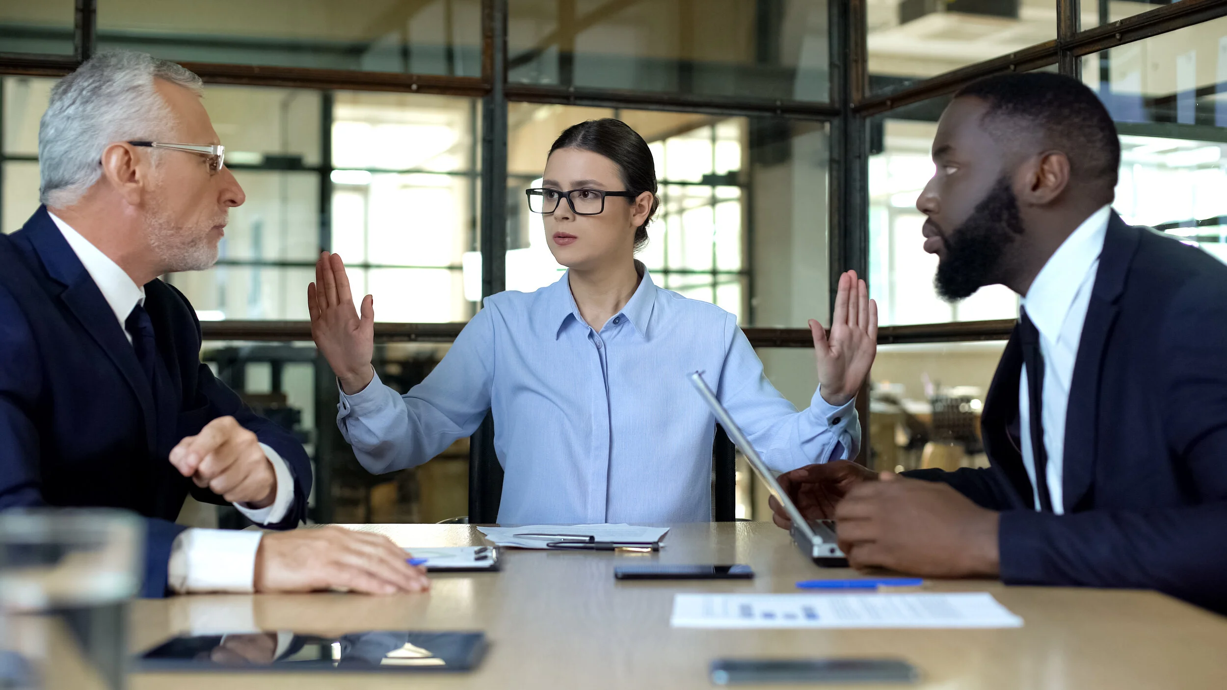 Three business professionals engaged in a serious discussion at a meeting, with a woman in the middle gesturing with her hands, situated in a modern office setting.