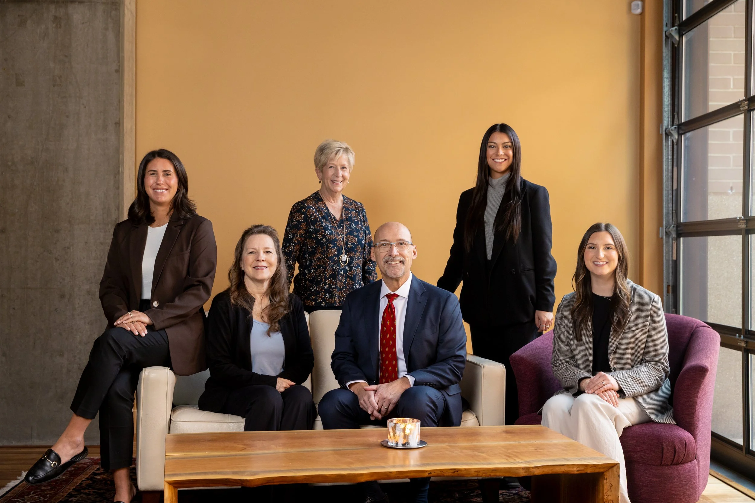 Group of seven people in a living room, including six women and one man, dressed in business attire, sitting and standing around a beige sofa and wooden coffee table, with a large window and yellow wall in the background.