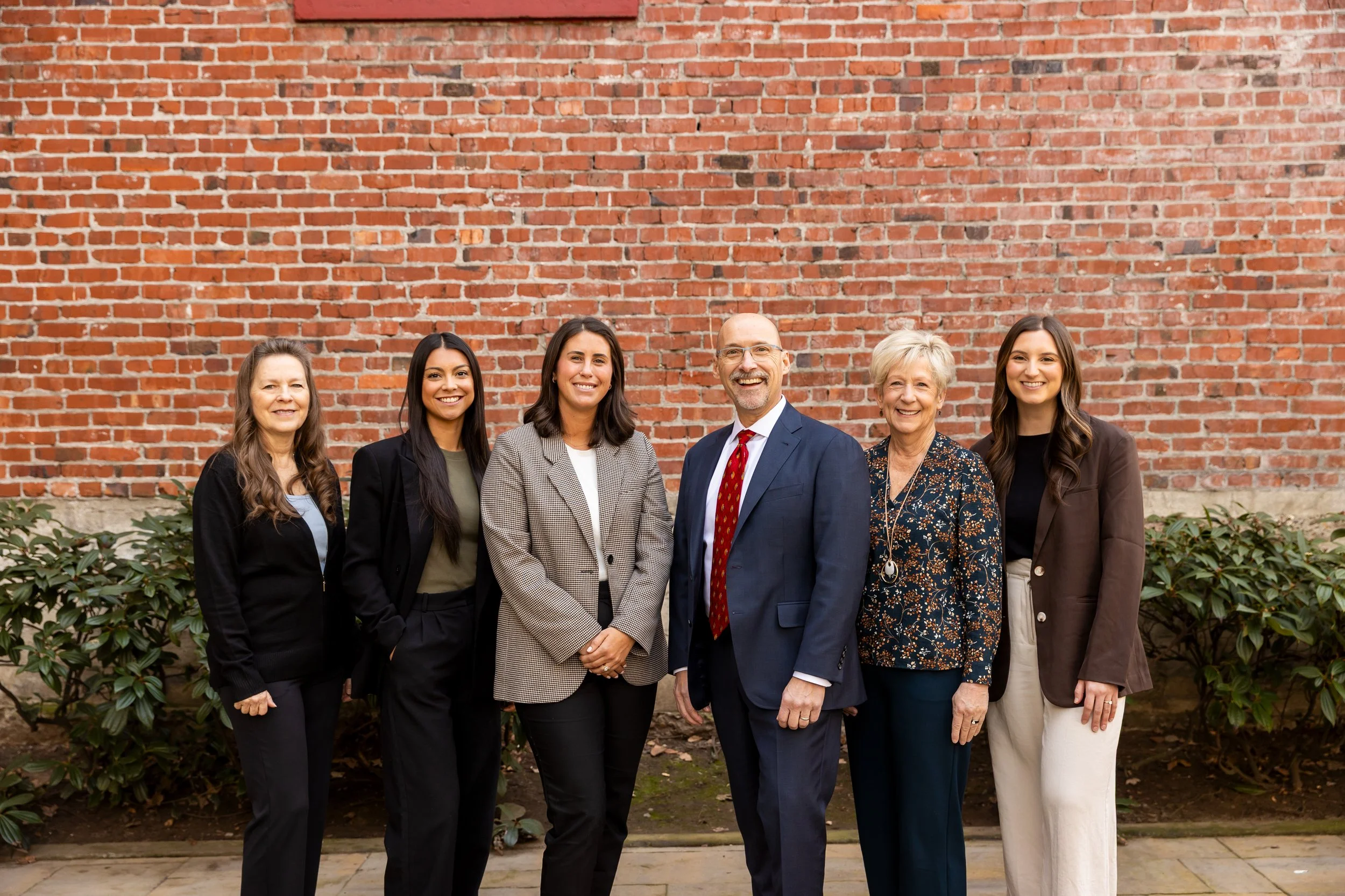A group of six professionally dressed people standing together outdoors in front of a brick wall and greenery, smiling at the camera.