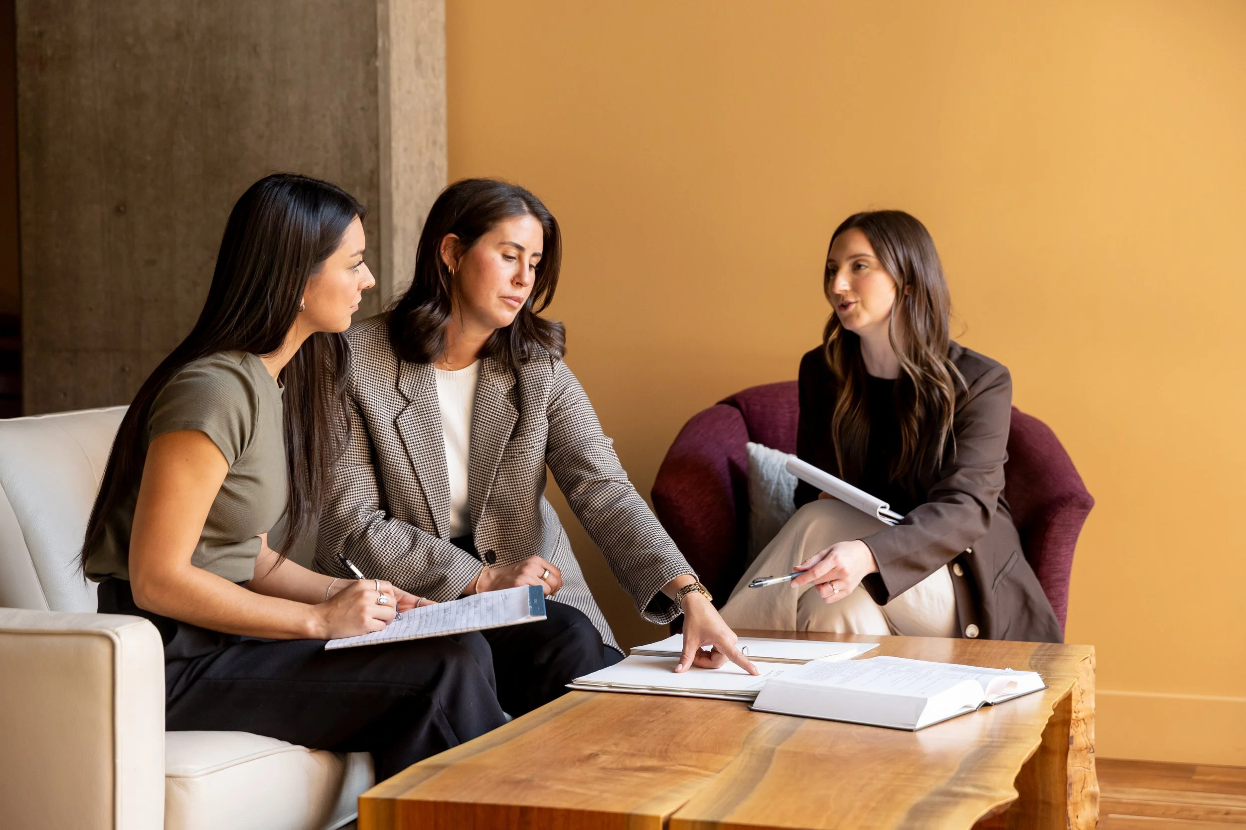 Three women engaged in a discussion during a meeting in a room with yellow walls. They are sitting on a beige sofa and a maroon armchair with documents and notebooks on a wooden table in front of them.