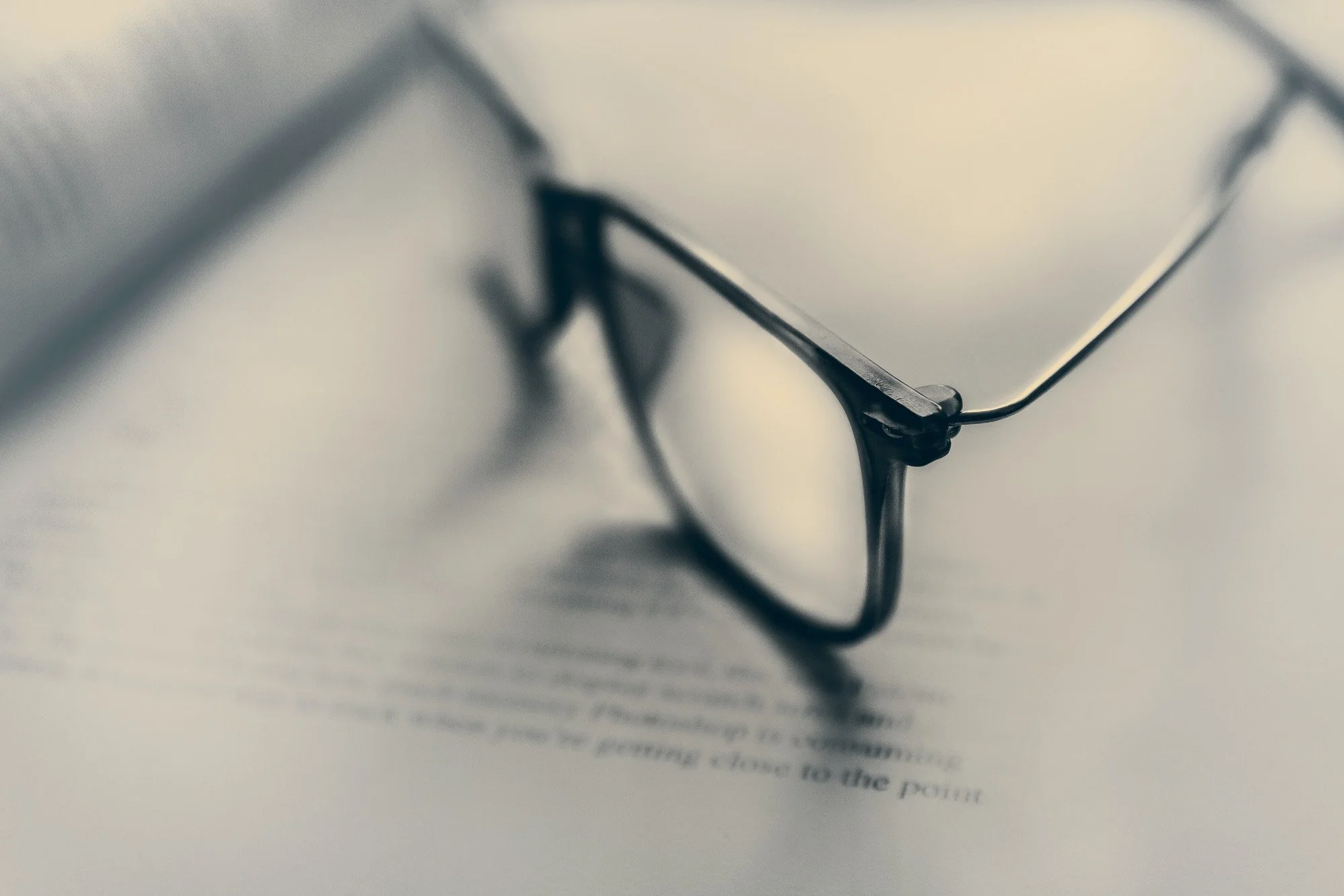 A close-up photograph of eyeglasses resting on top of a printed document or book page, with the text blurred and out of focus.