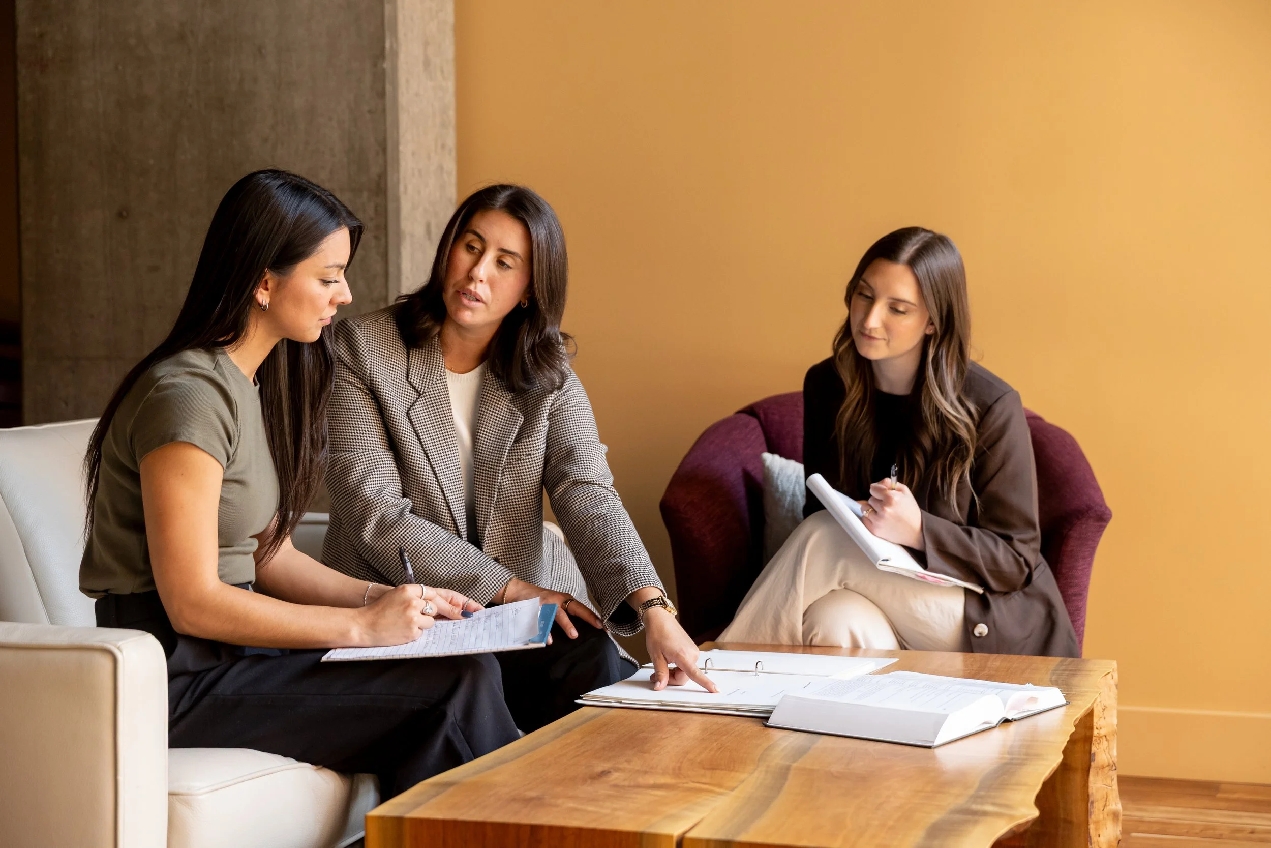 Three women sitting on a sofa and a chair around a wooden table, engaged in a discussion, with some papers and notebooks in front of them.