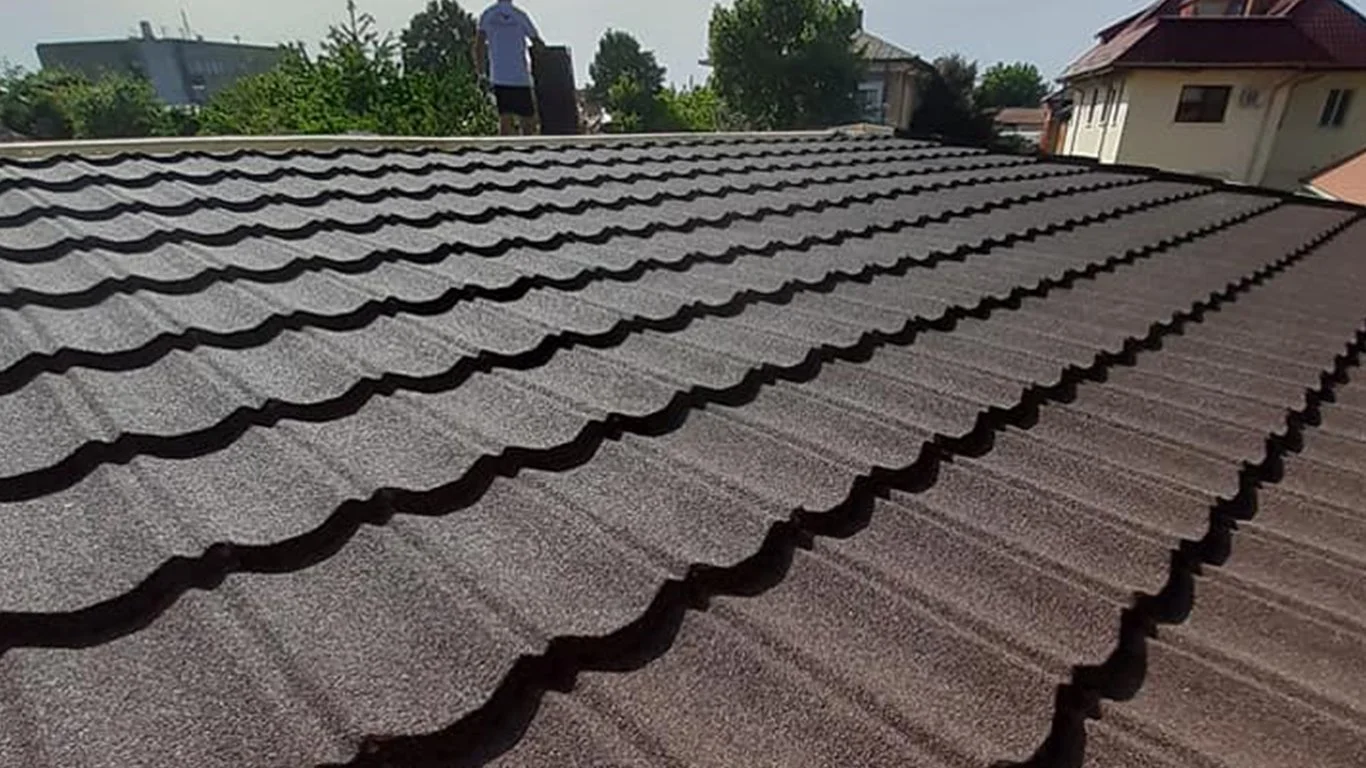 Close-up view of a tiled roof with dark shingles, with a person standing on the roof in the background, and trees and houses visible beyond.