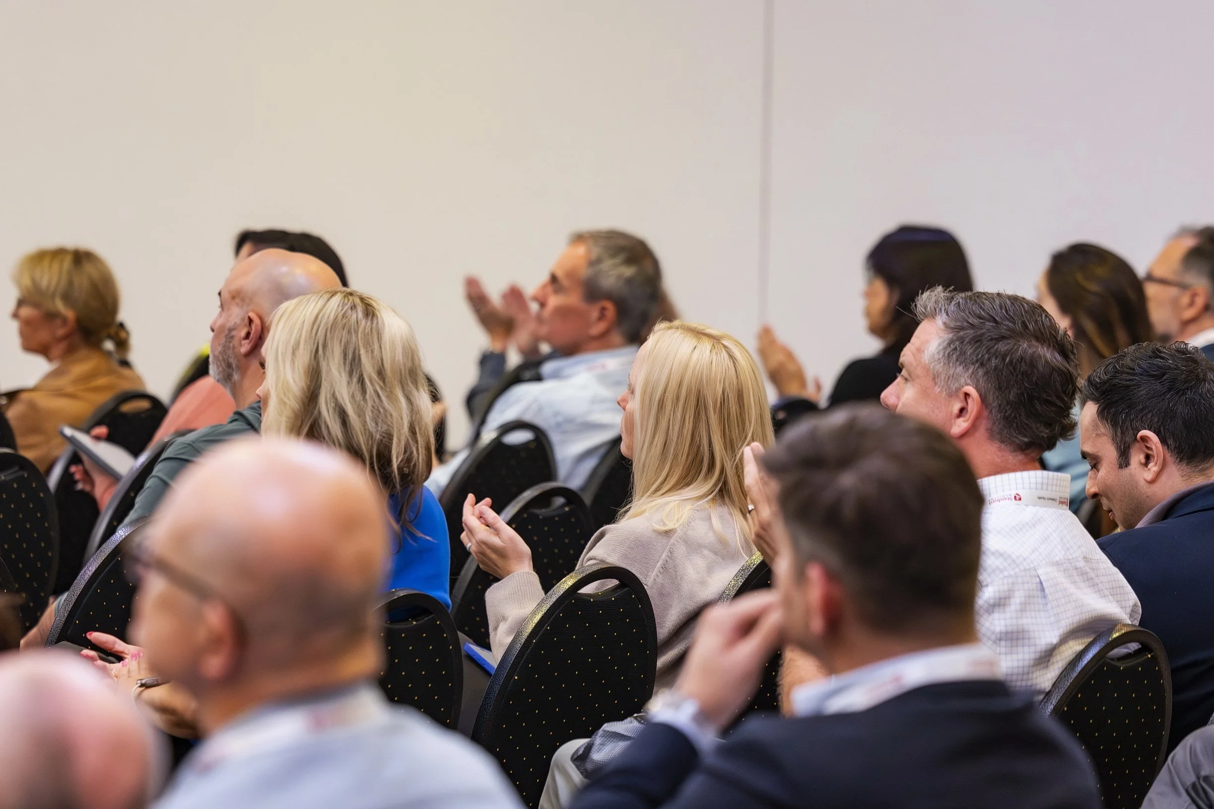 Audience seated and listening at a conference or seminar, with diverse men and women facing forward, some taking notes or clapping.