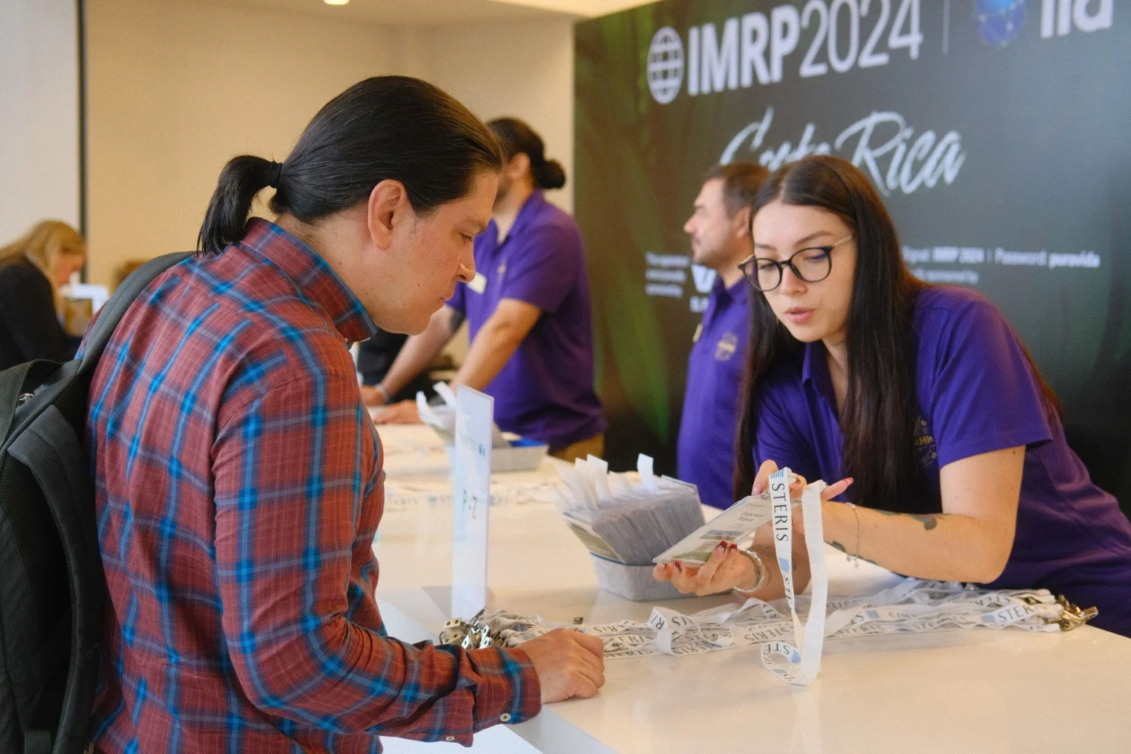 A woman with long dark hair and glasses checks in with a staff member at a registration desk, while a man with a backpack looks on. The staff member is handing out informational materials. In the background, there are other staff members and a large 