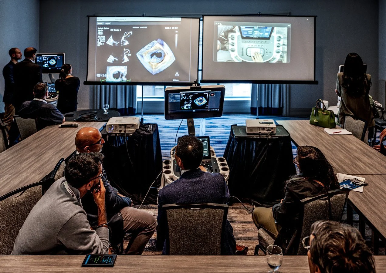 Medical professionals and conference attendees observing ultrasound imaging and medical equipment during a presentation in a conference room.