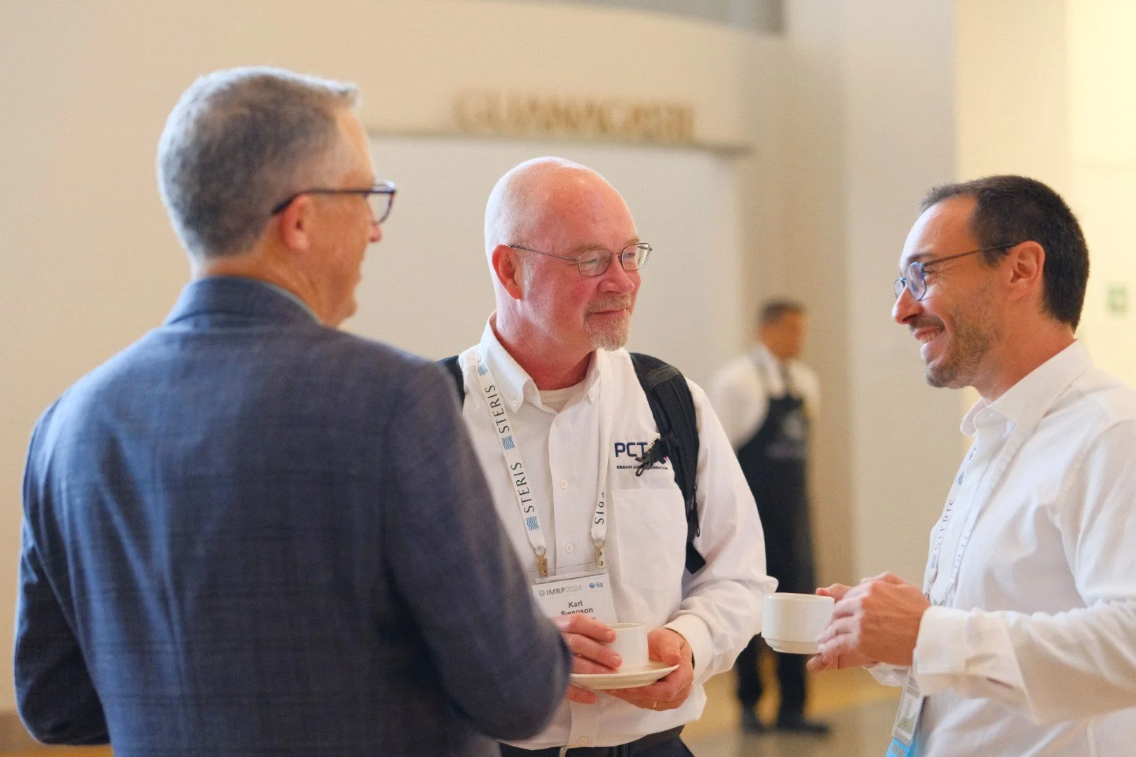 Three men engaged in conversation at a professional event, with one holding a coffee cup, in a well-lit room with a blurred background.