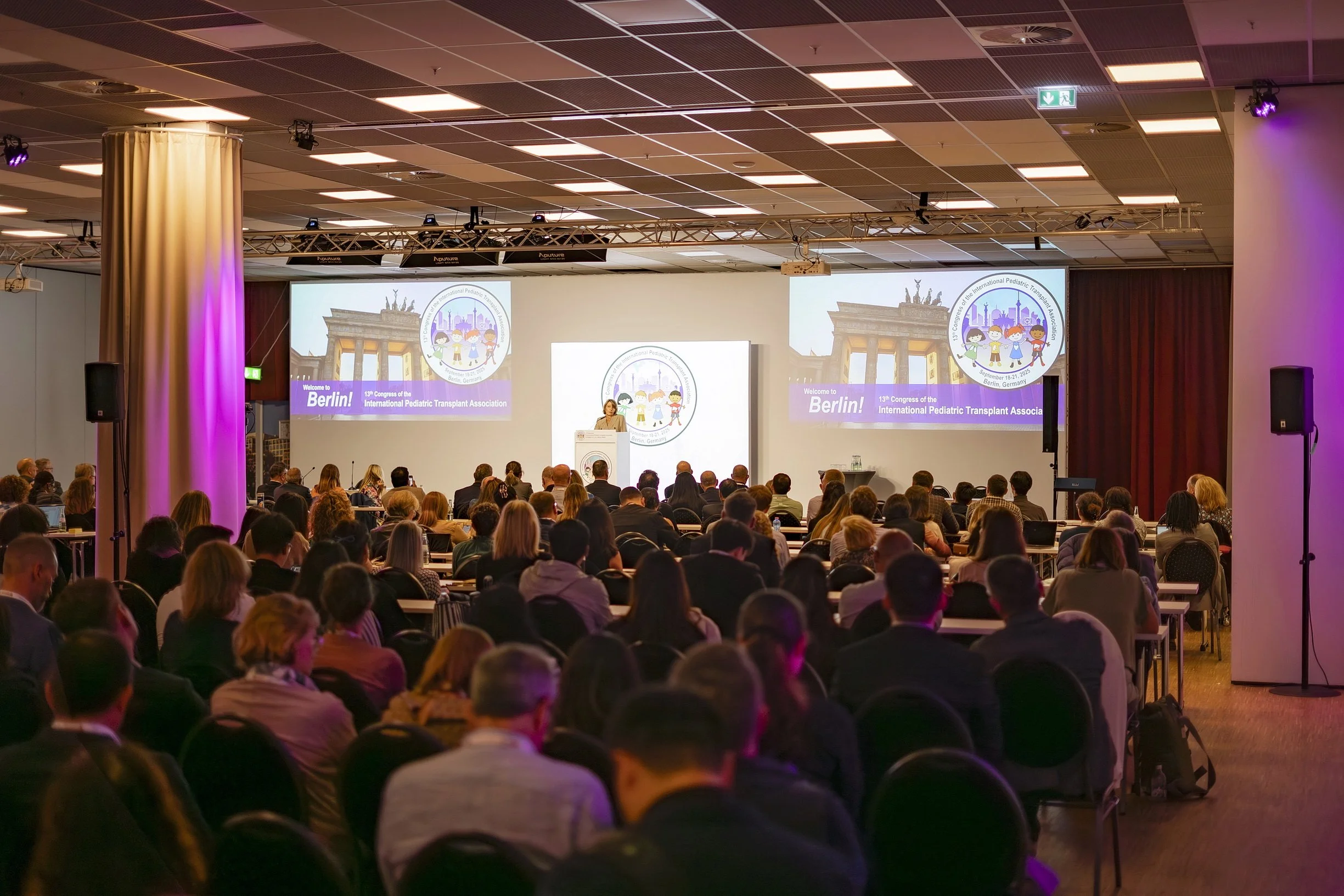 Conference room filled with attendees listening to a speaker at the podium on stage. The stage has large screens displaying logos and text about Berlin and the International Pediatric Transplant Association.