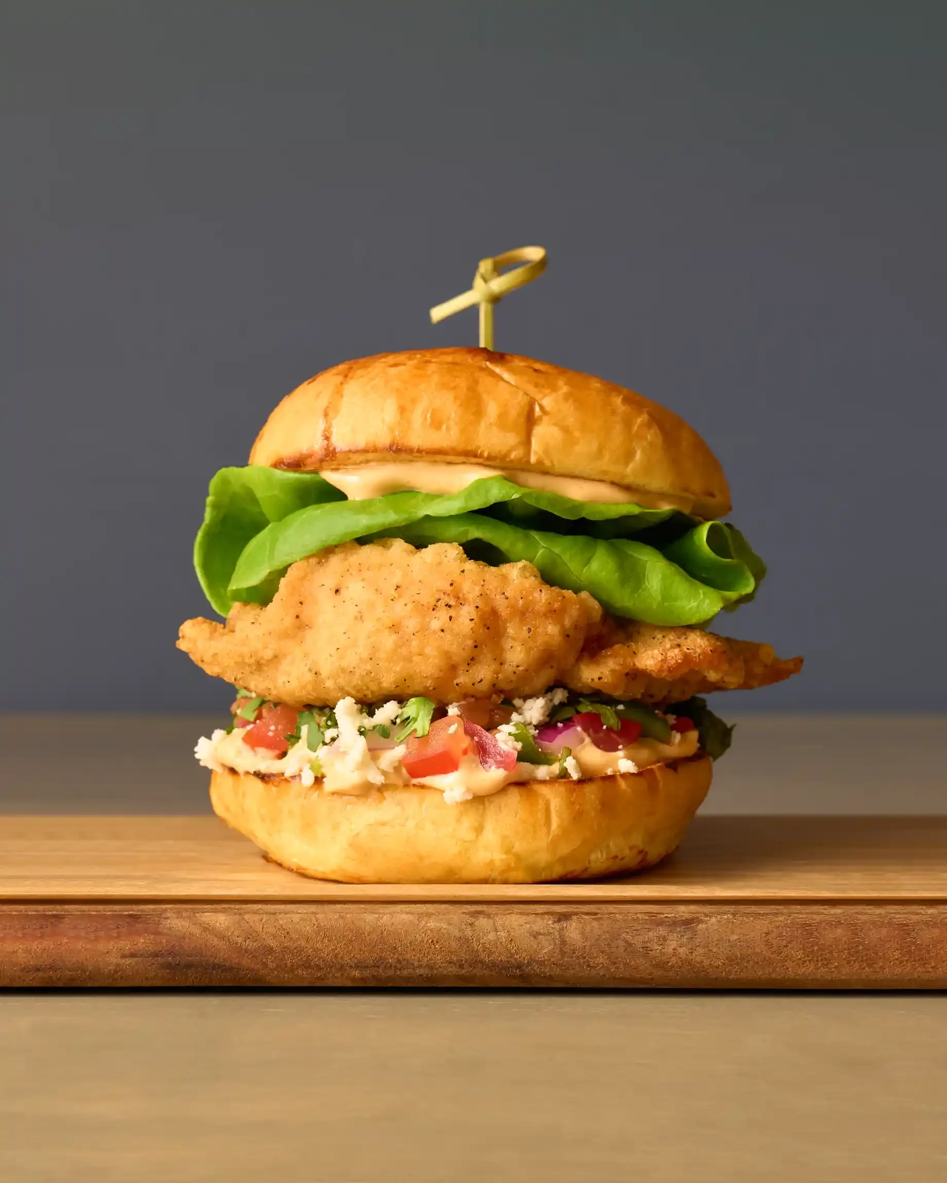 A fried chicken sandwich with lettuce, tomato, cheese, and sauce in a brioche bun on a wooden board. By Doylestown, PA food photographer & food stylist team Bartholomew Studio.