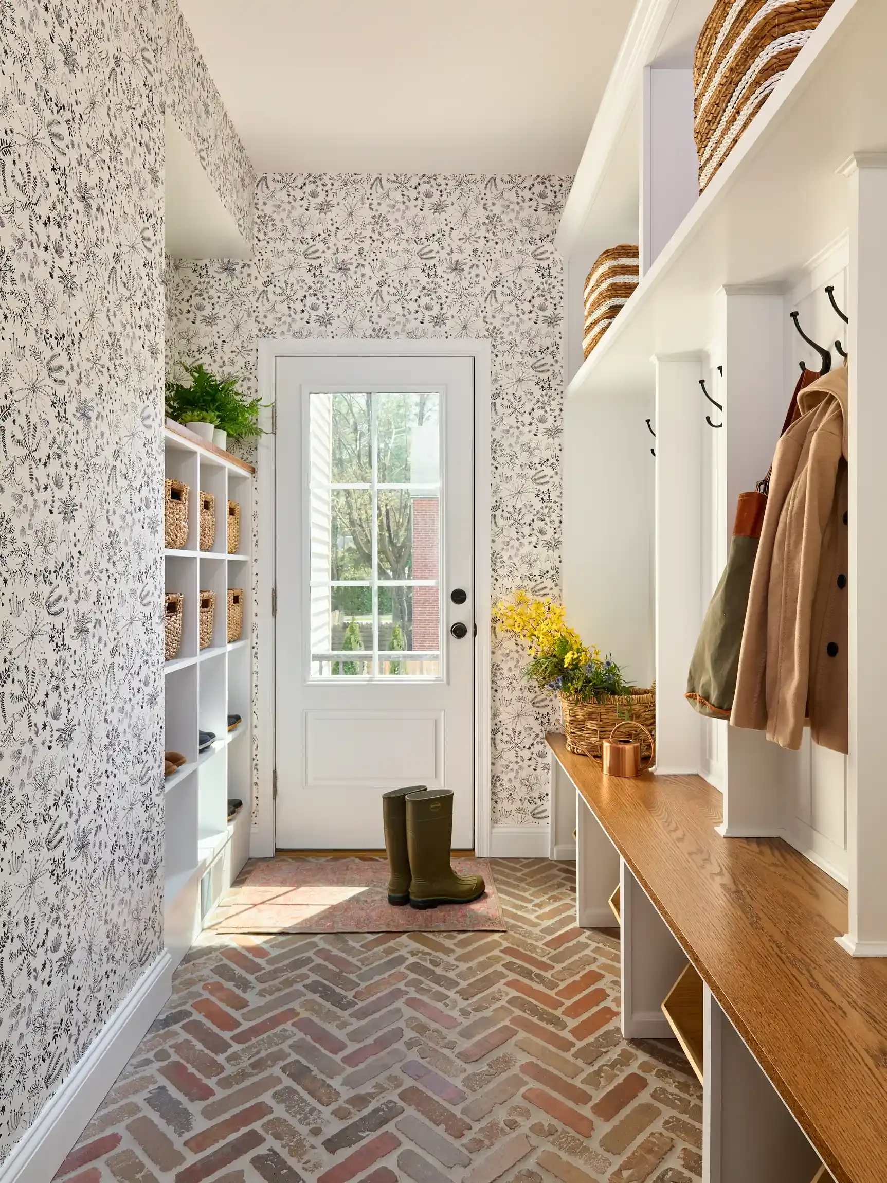 Entryway with floral wallpaper, a white door with glass window, green rain boots on a small rug, shelves with baskets, a wooden bench and coat hooks with jackets hanging. By New Jersey interior photographer Bartholomew Studio.