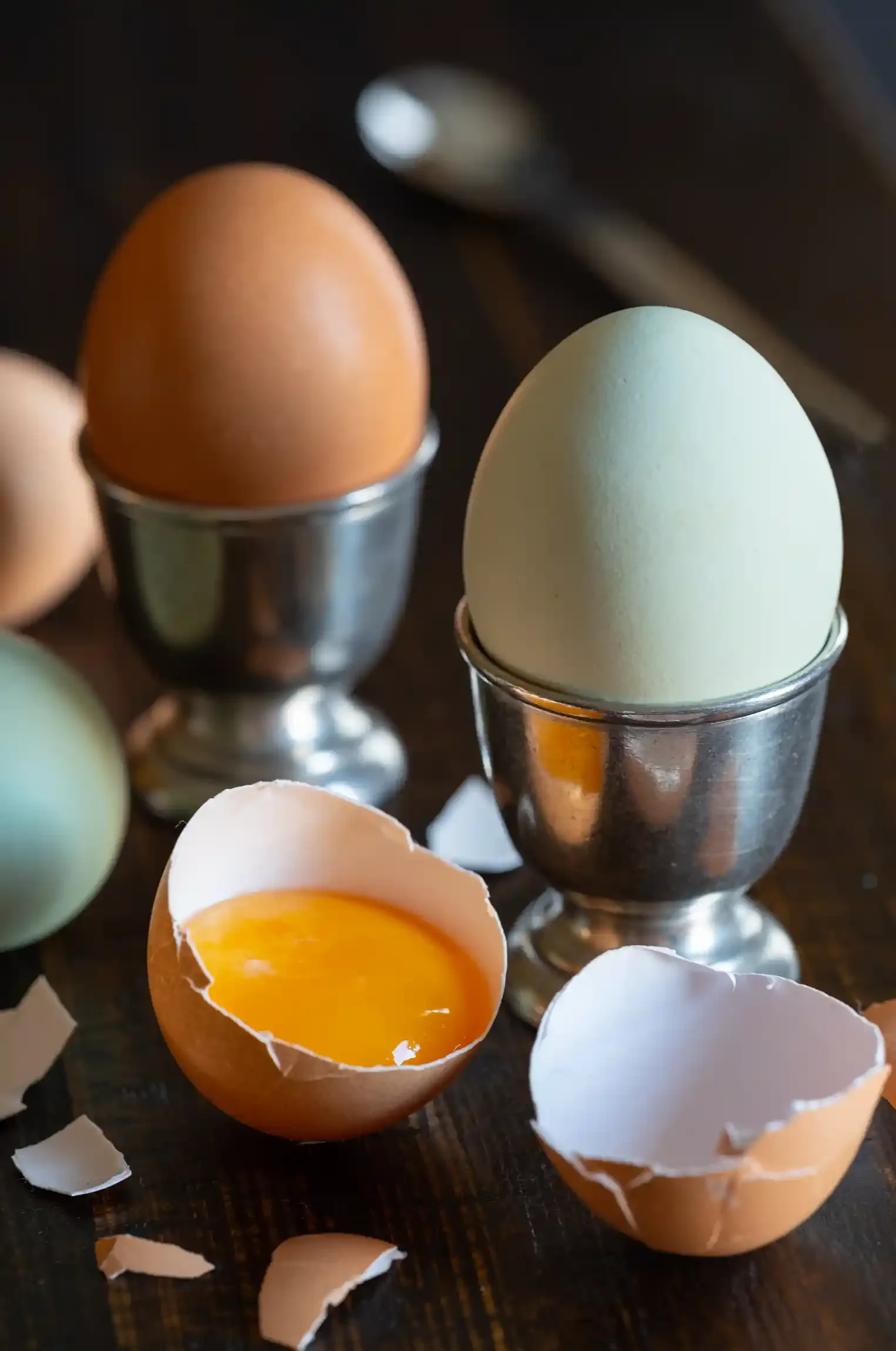 Two eggs, one brown and one light green, in metal egg cups on a dark wooden surface. One egg is cracked open, revealing yolk and broken eggshell pieces, while the other egg remains intact. By PA & NJ food photographer & food stylist tea