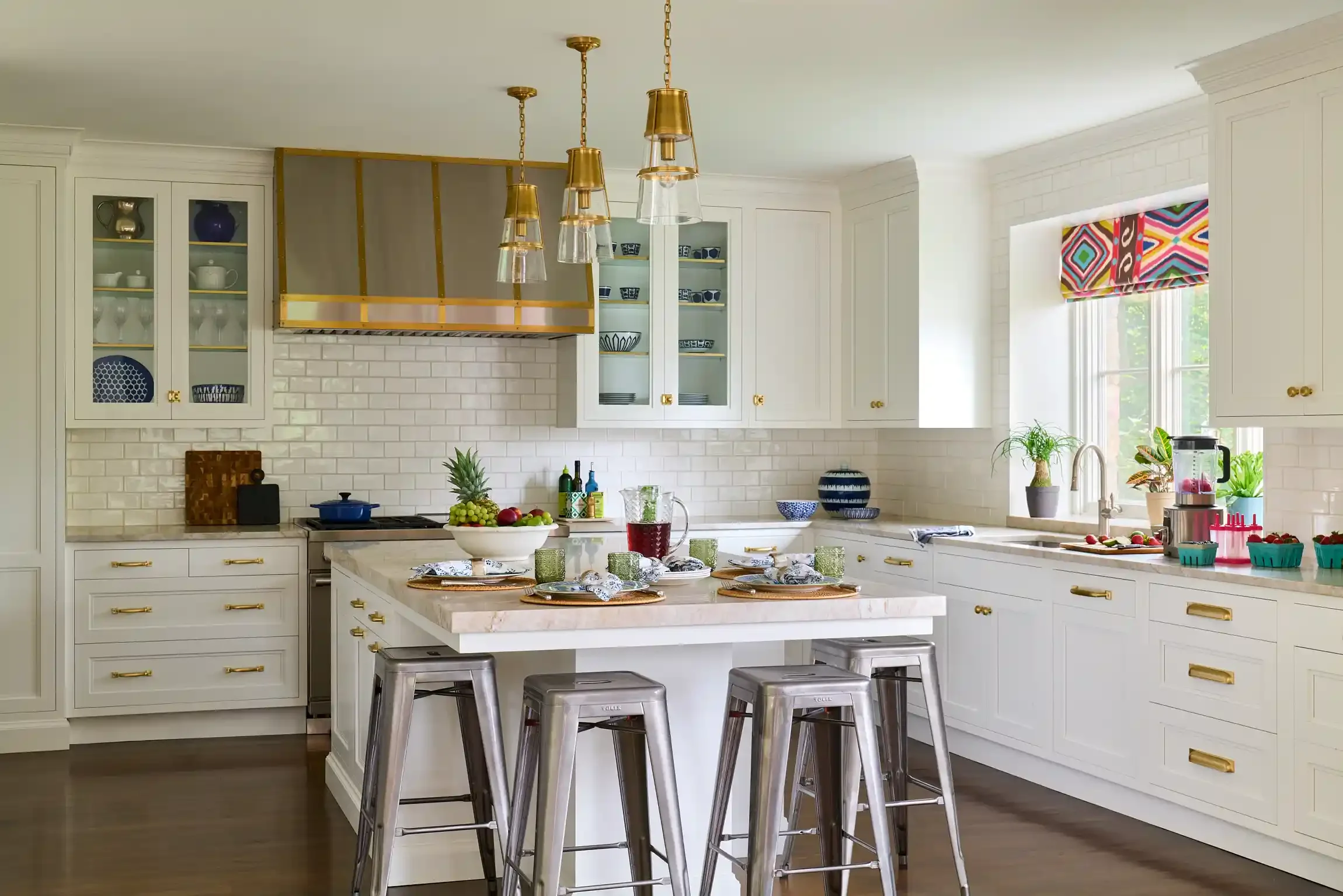 Modern white kitchen with gold accents, kitchen island with stools, hanging pendant lights, window with colorful Roman shade, open cabinetry, and various kitchen accessories. By New Jersey interior photographer Bartholomew Studio.
