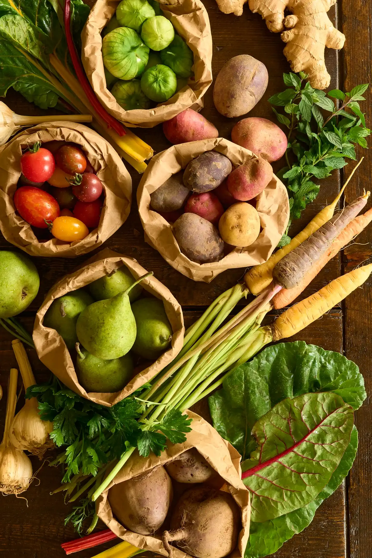 Fresh vegetables including tomatoes, potatoes, carrots, ginger, garlic, leafy greens, and herbs arranged on a wooden surface. By Philadelphia, PA & NJ food photographer & food stylist team Bartholomew Studio.