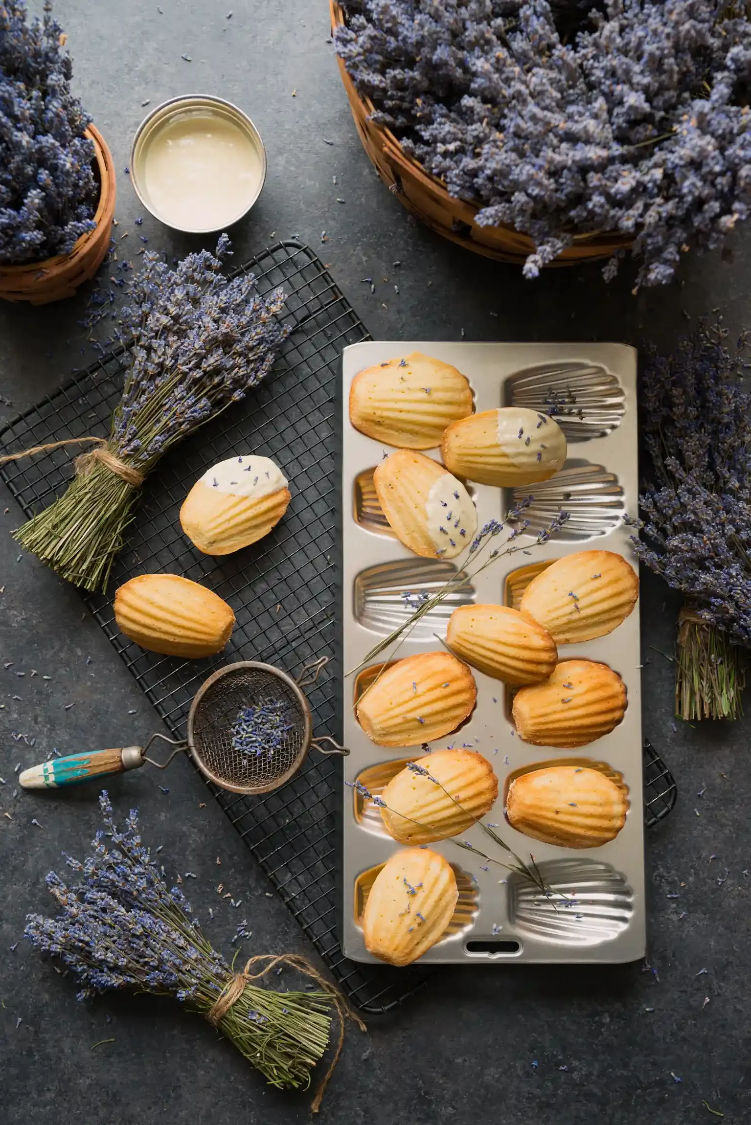 French madeleines with dried lavender flowers, some scattered on a gray surface and surrounded by lavender bunches. By Lehigh Valley & Bucks County, PA food photographer & food stylist team Bartholomew Studio.