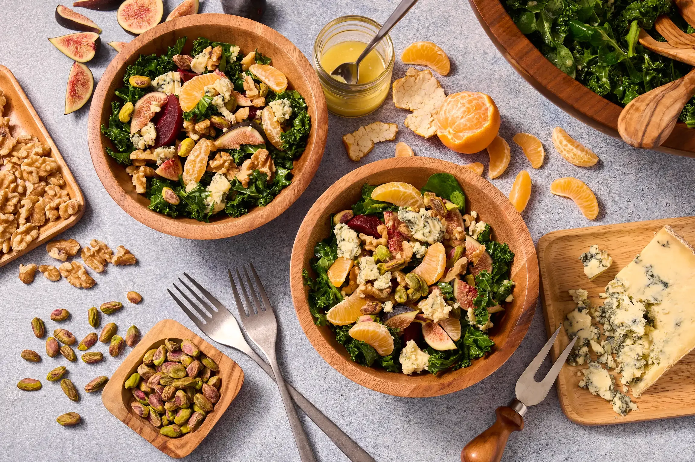 Mixed salads with figs, oranges, walnuts, blue cheese, and pistachios, along with scattered nuts and fruit on a gray surface.