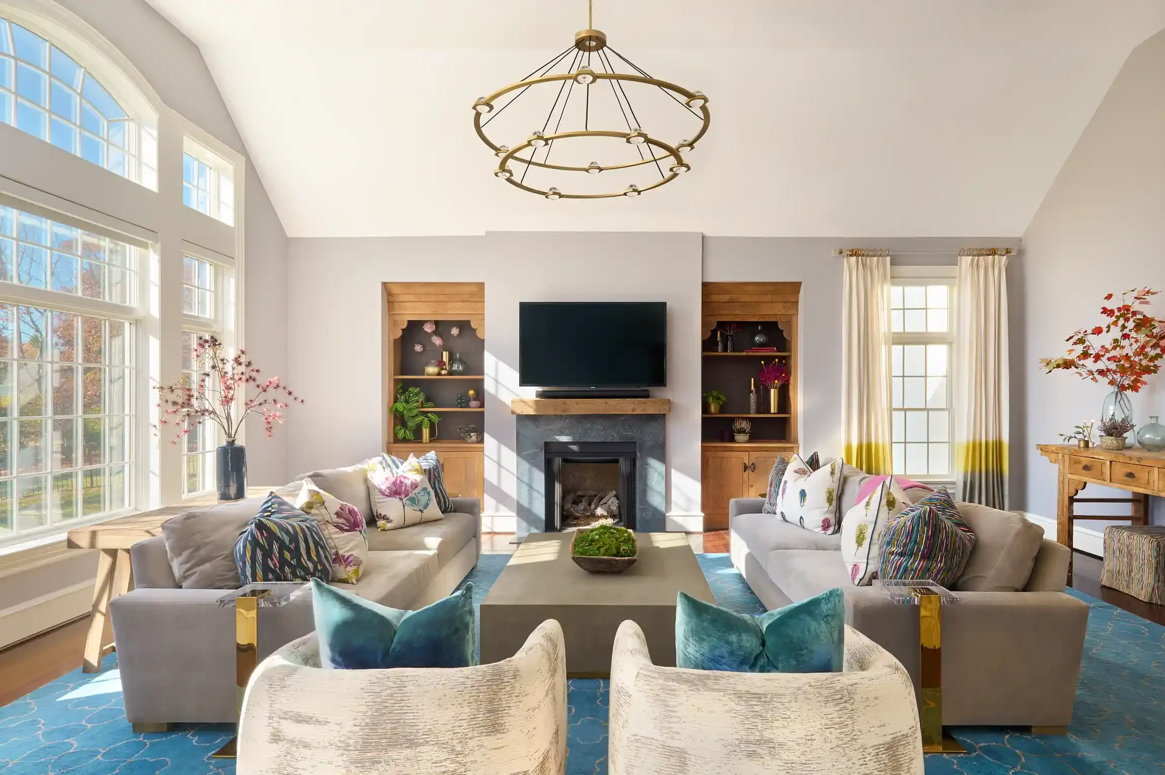 Bright living room with large windows, modern sofa, armchairs, coffee table, fireplace with mounted TV, decorative built-in shelves, chandelier, and colorful cushions. By Philadelphia, PA area interior photographer.