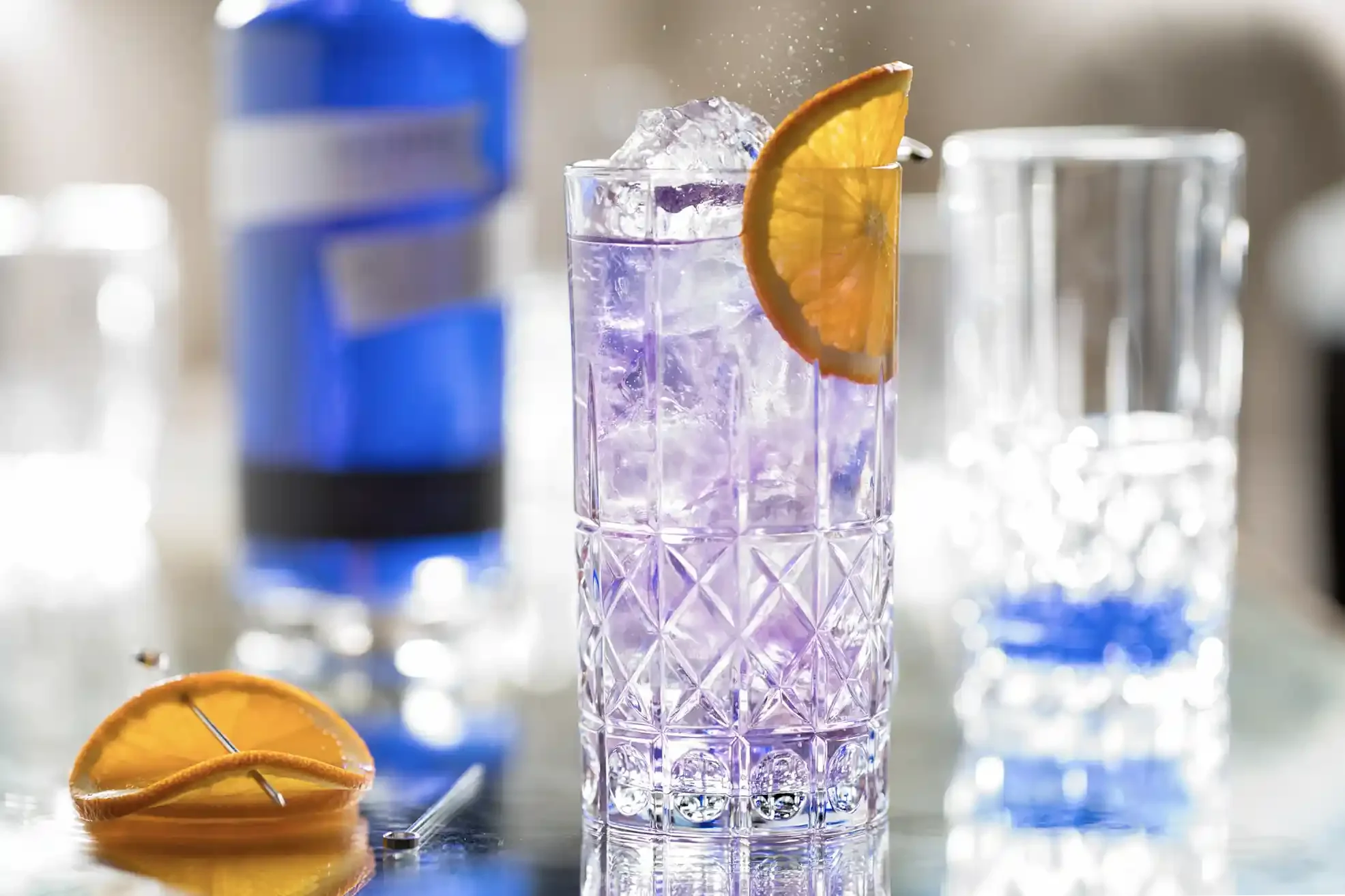 A tall glass of a purple floral gin & tonic cocktail with ice, garnished with an orange slice, on a reflective surface with two empty glasses and a blurred bottle in the background. By Philadelphia, PA, NJ drink photographer & drink stylist team.