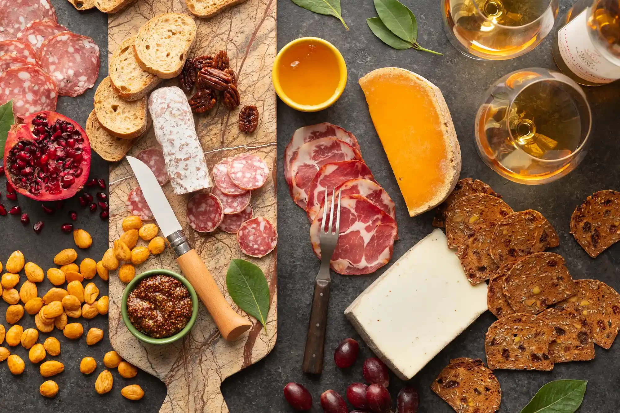 A charcuterie board with slices of different cured meats, cheeses, crackers, grapes, pomegranate, nuts, and dips, accompanied by glasses of white wine. By Philadelphia, PA food photographer & food stylist team Bartholomew Studio.