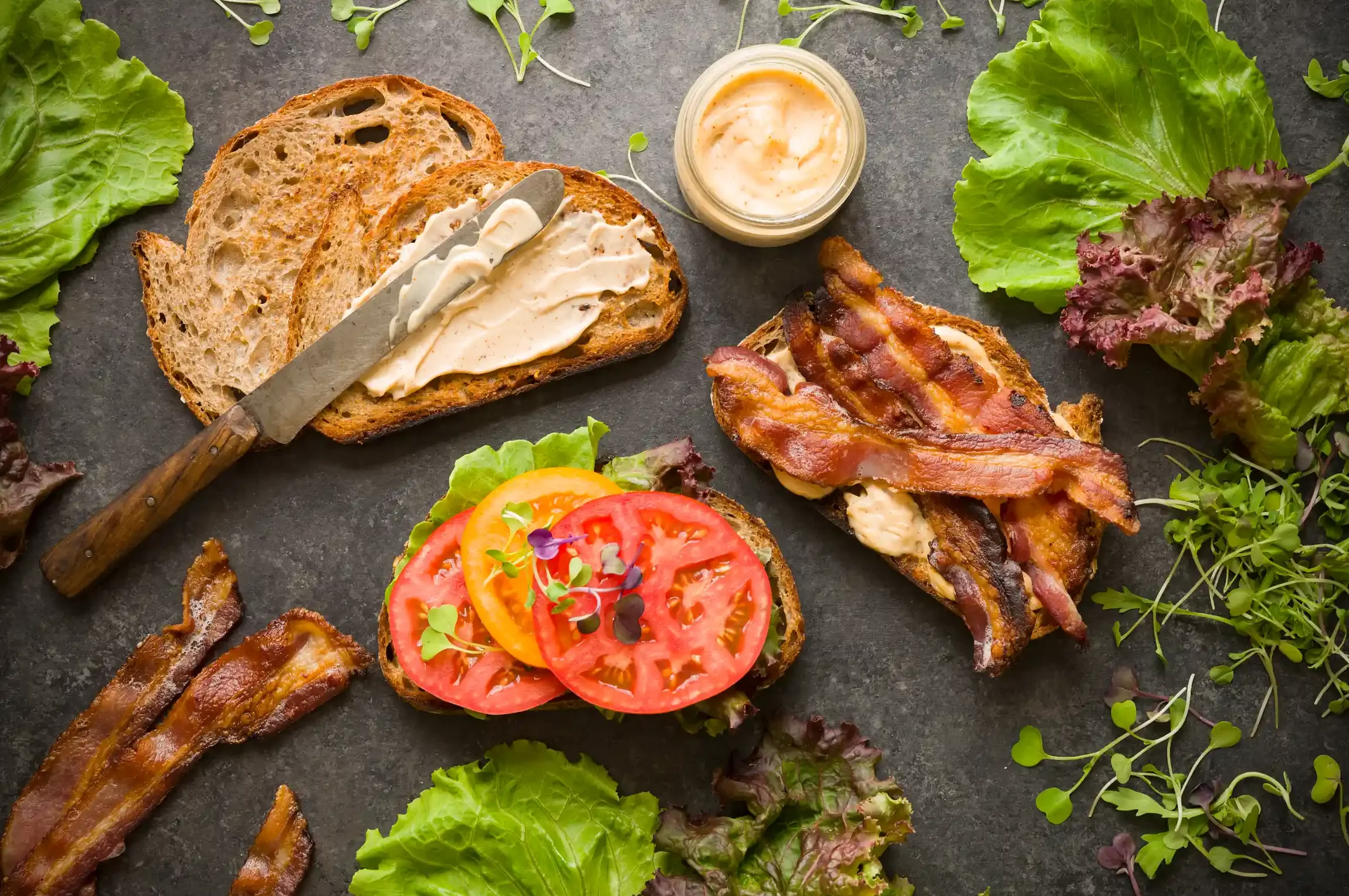 Open-faced BLT style sandwich with tomatoes, bacon, lettuce, and microgreens on dark surface with slices of bread, bacon, and a small bowl of sauce. By Bucks County, PA & NJ food photographer & food stylist team Bartholomew Studio.