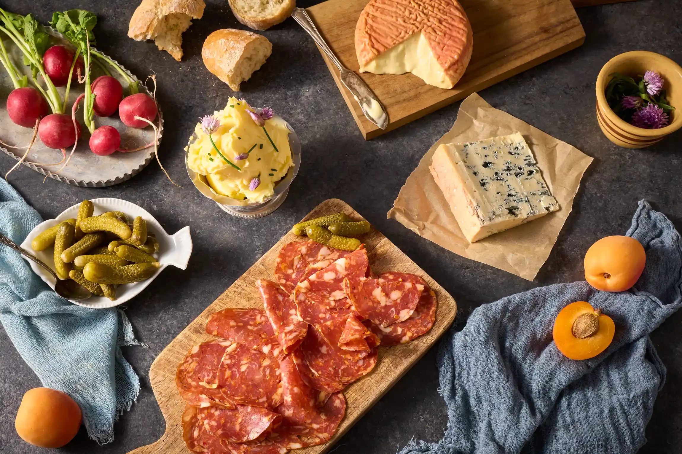 Assorted cheese, cured meats, pickles, radishes, apricots with flowers, bread pieces, and small flowers on a dark table for a charcuterie spread. By Philadelphia, PA food photographer & food stylist team Bartholomew Studio.