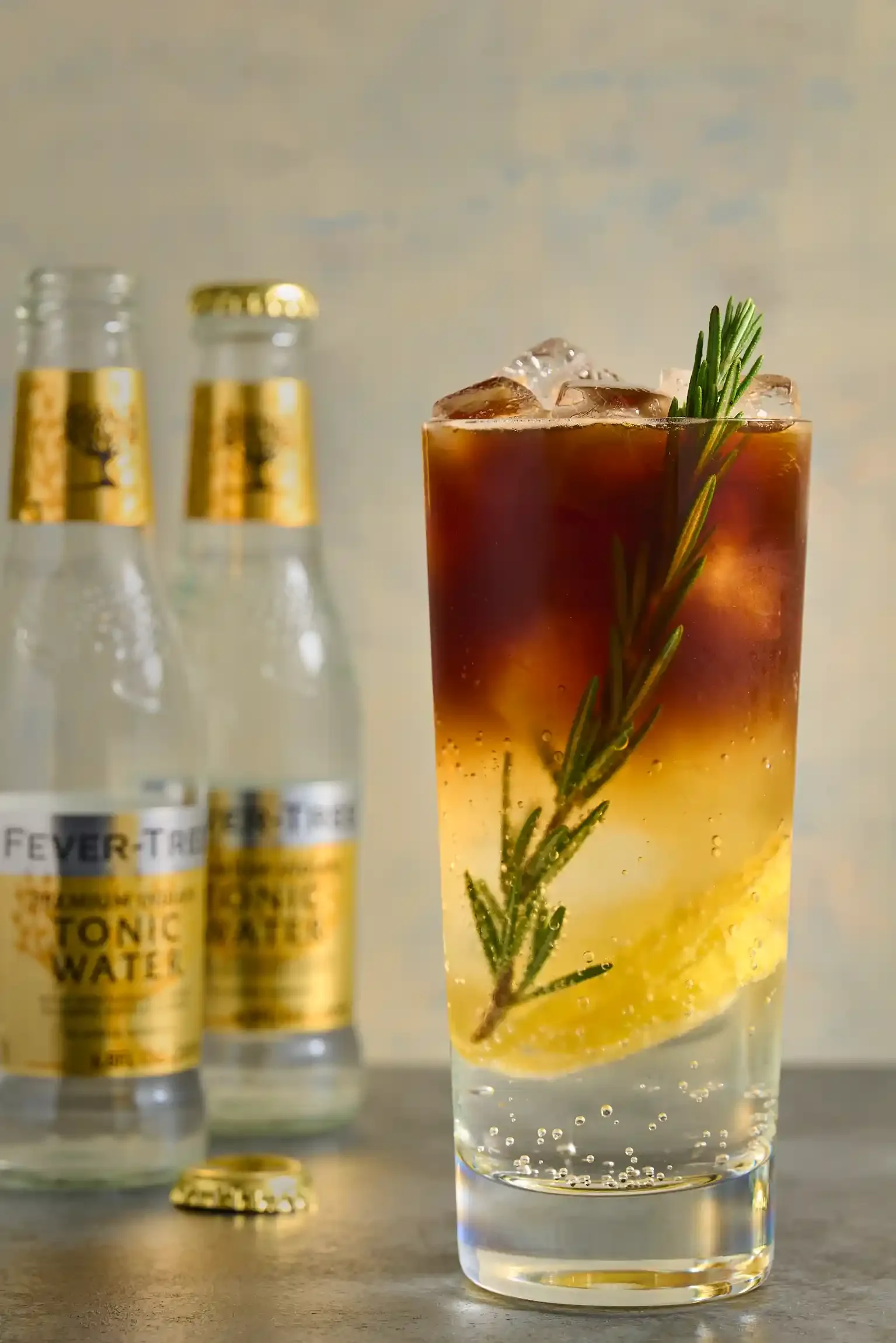 A tall glass of iced soda with a sprig of rosemary and lemon inside, with two bottles of tonic water in the background. By Philadelphia, PA & NJ drink photographer & drink stylist team Bartholomew Studio.